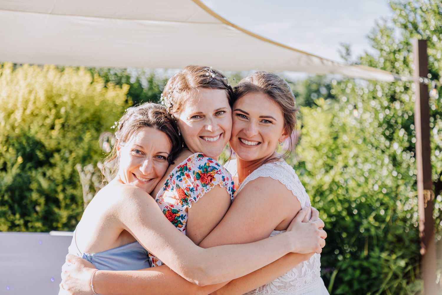 Three women in wedding attire share joyful group hug under outdoor canopy.