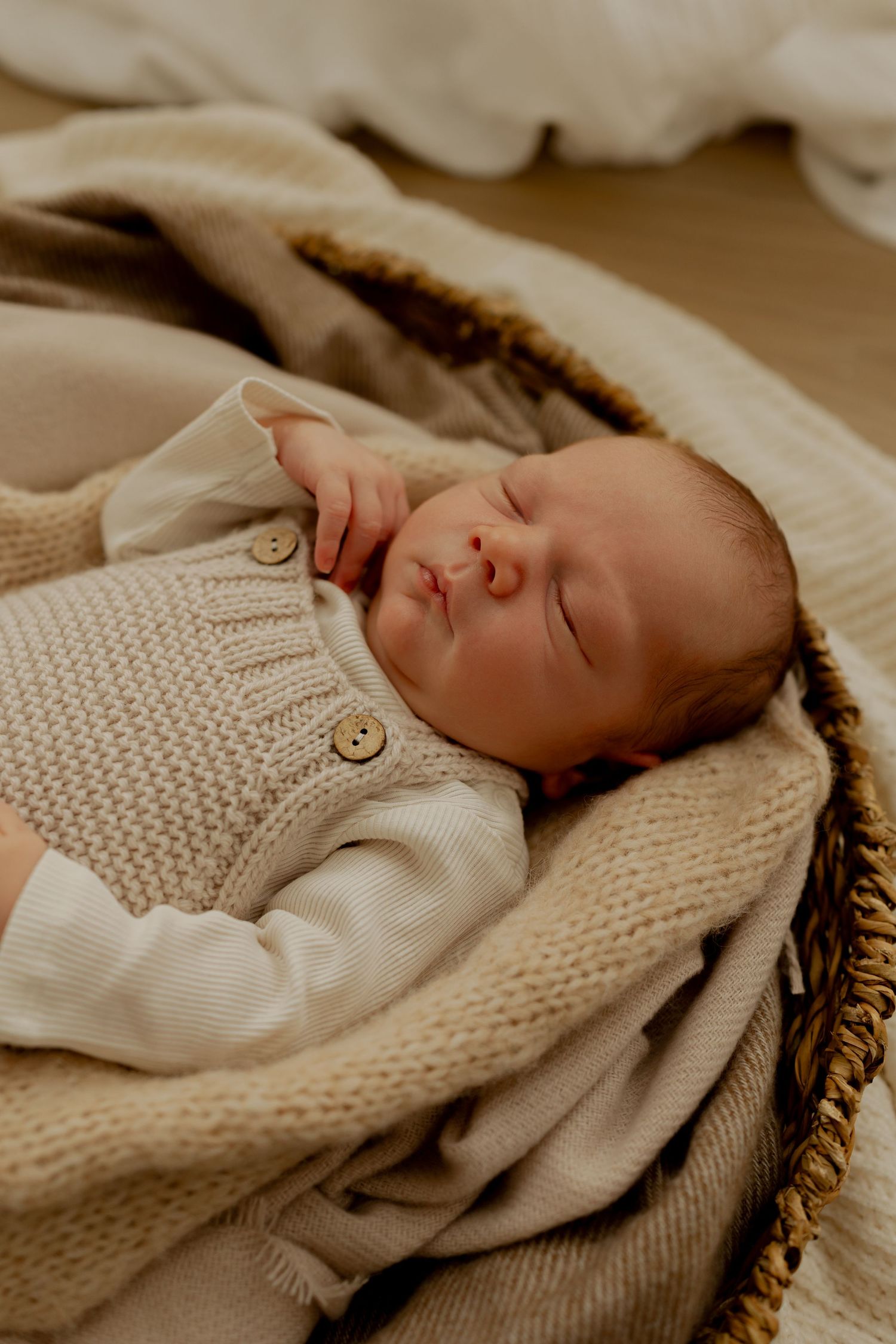 Peaceful newborn baby sleeping in a wicker basket wrapped in soft beige knit blankets wearing a cream sweater onesie.