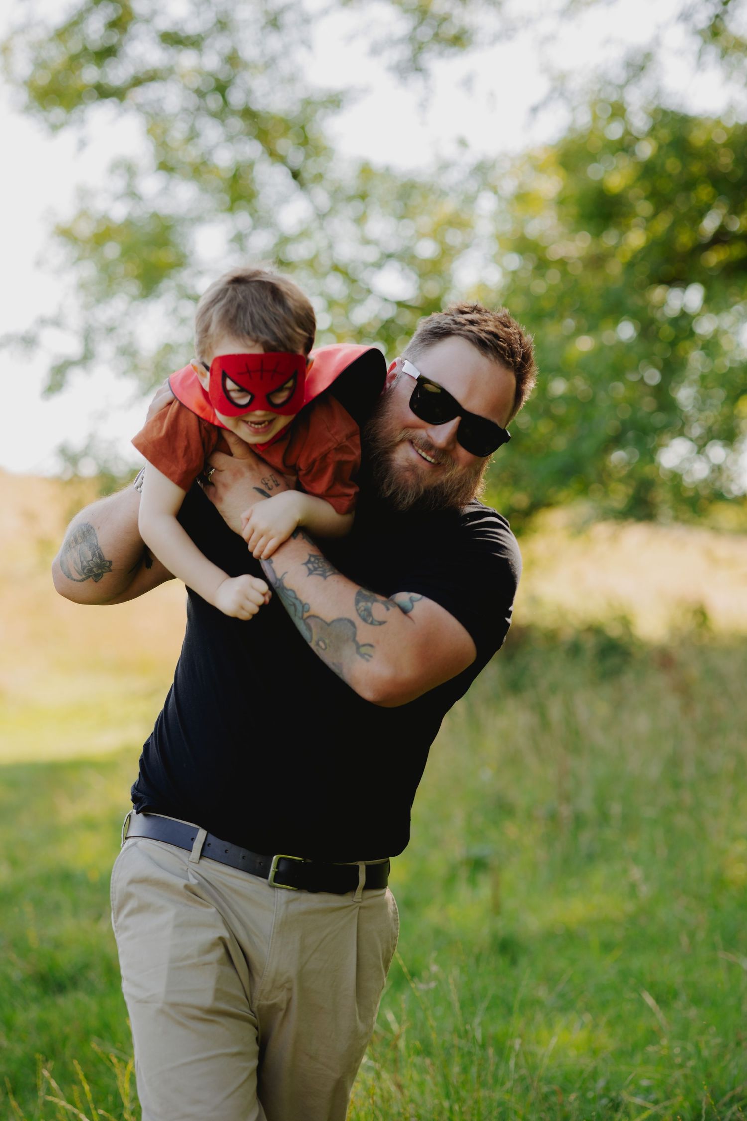 Adult holding child wearing Spider-Man mask in sunny outdoor setting.