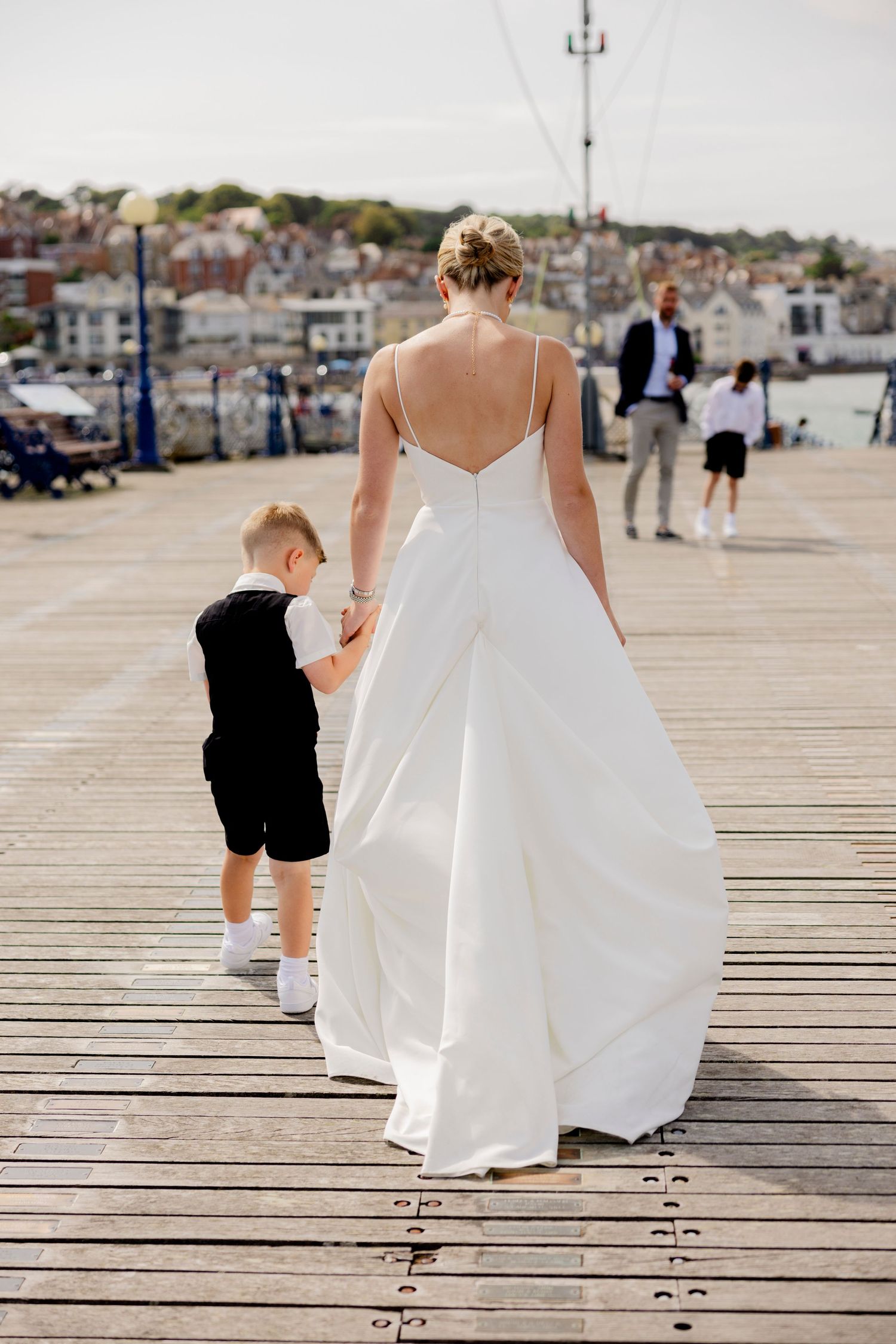 Bride in flowing white dress walking with young ring bearer on wooden pier.
