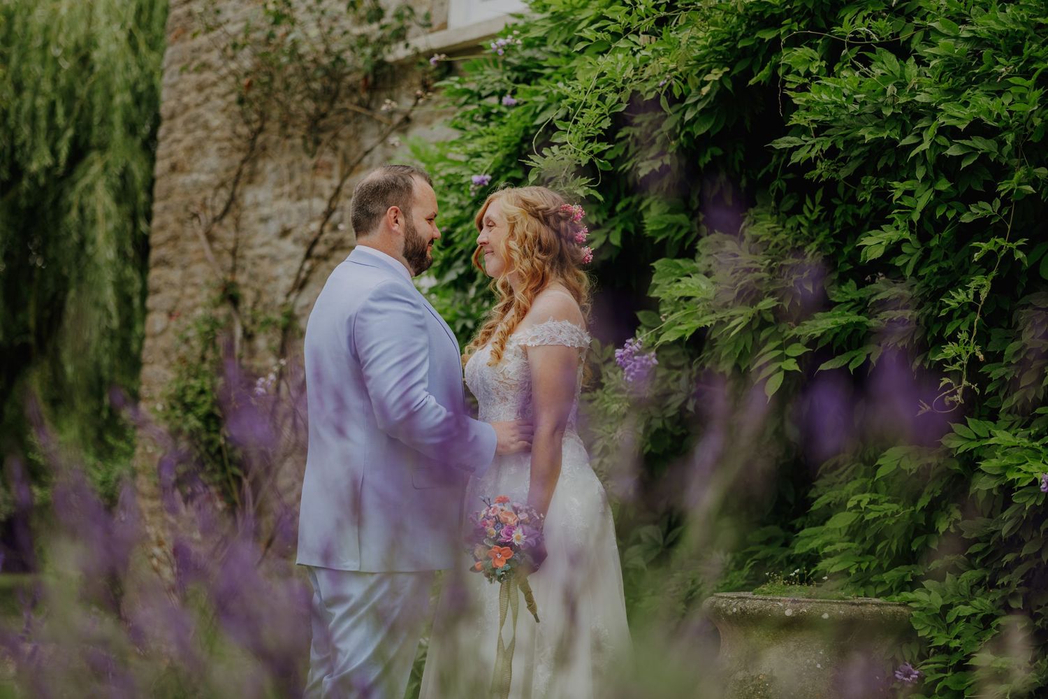 Romantic wedding couple embraces among purple flowers and greenery.