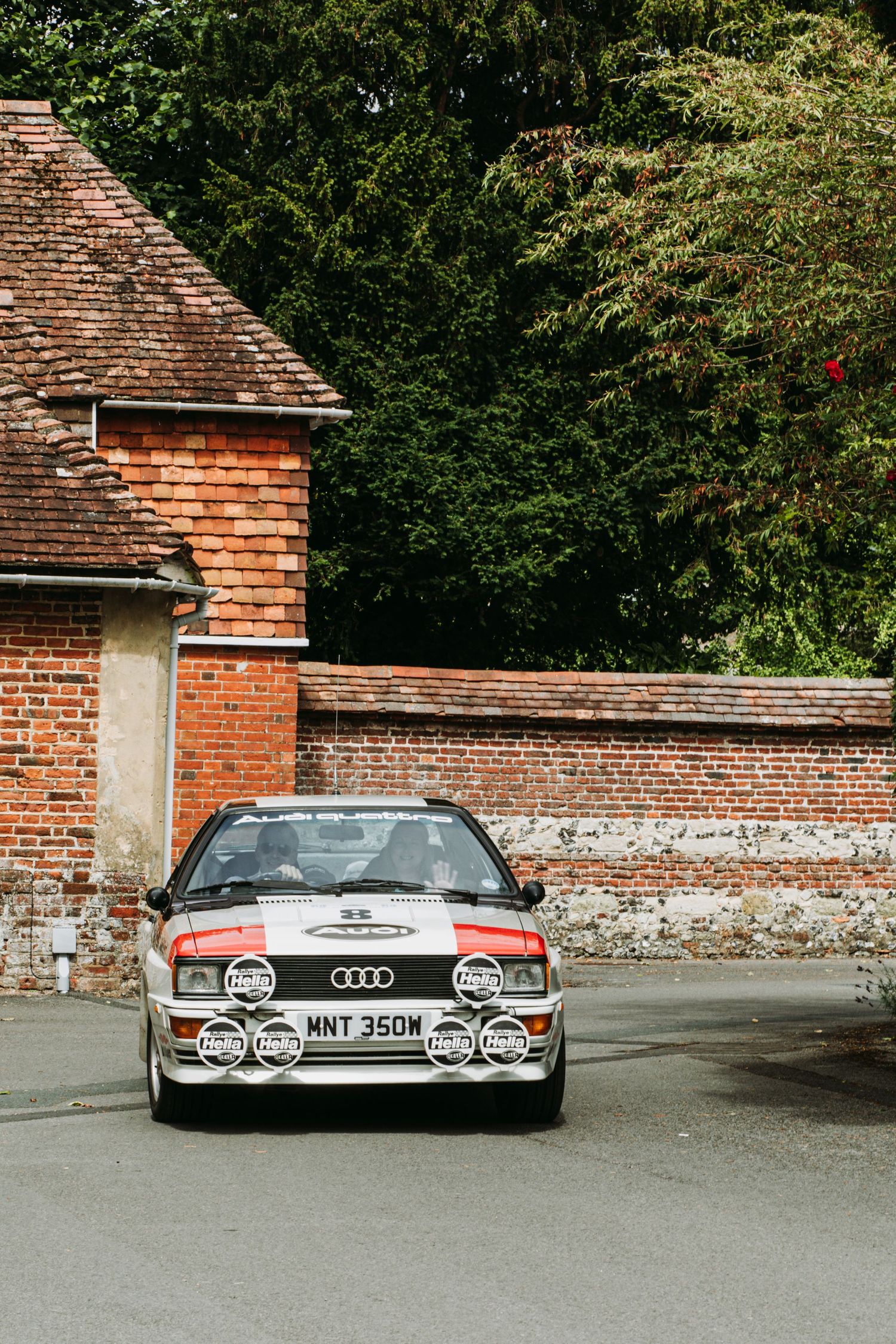 Vintage Audi Quattro with rally lights parked against brick wall and trees.
