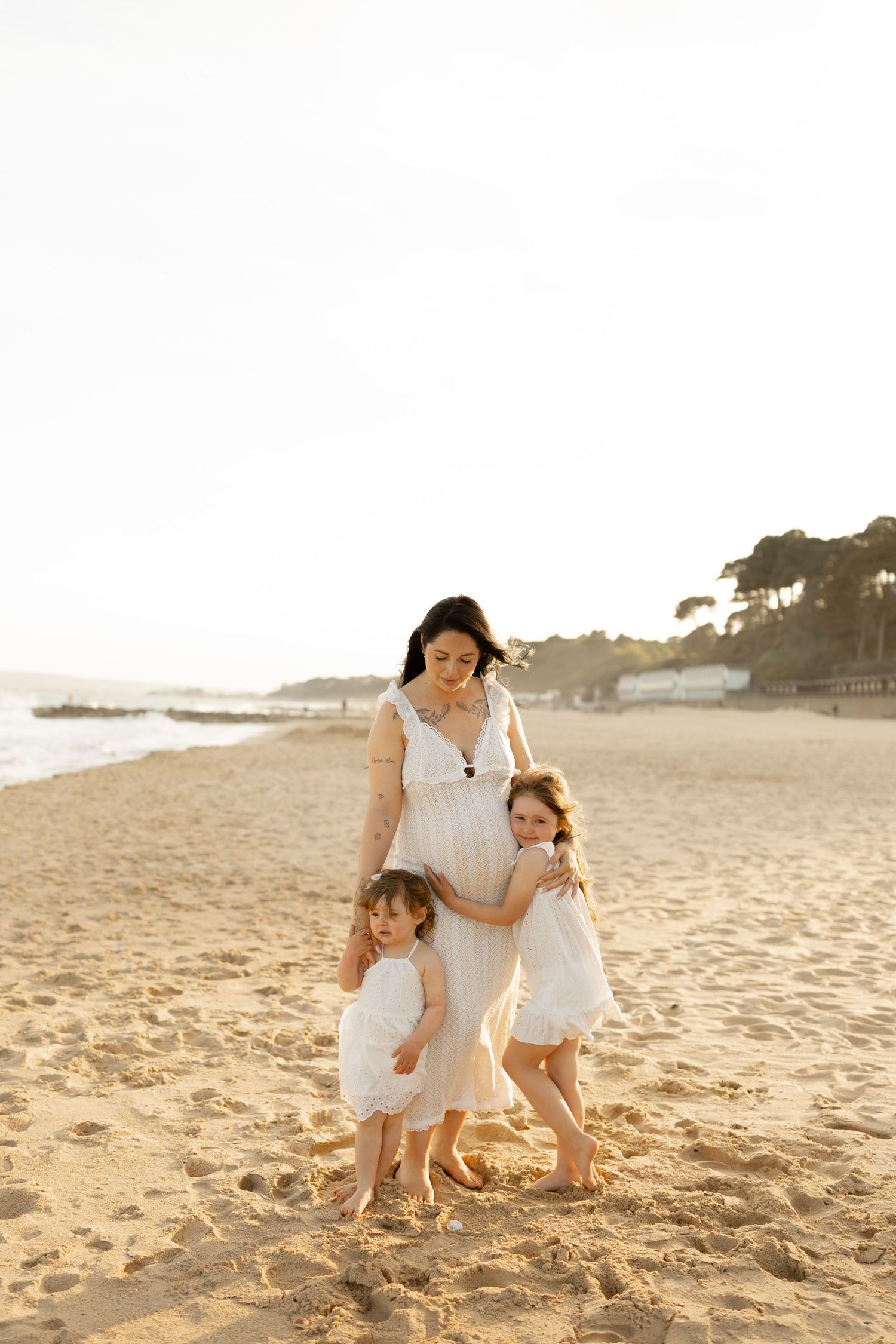 Pregnant woman in white dress with two young daughters in white dresses on a sunny beach at golden hour.