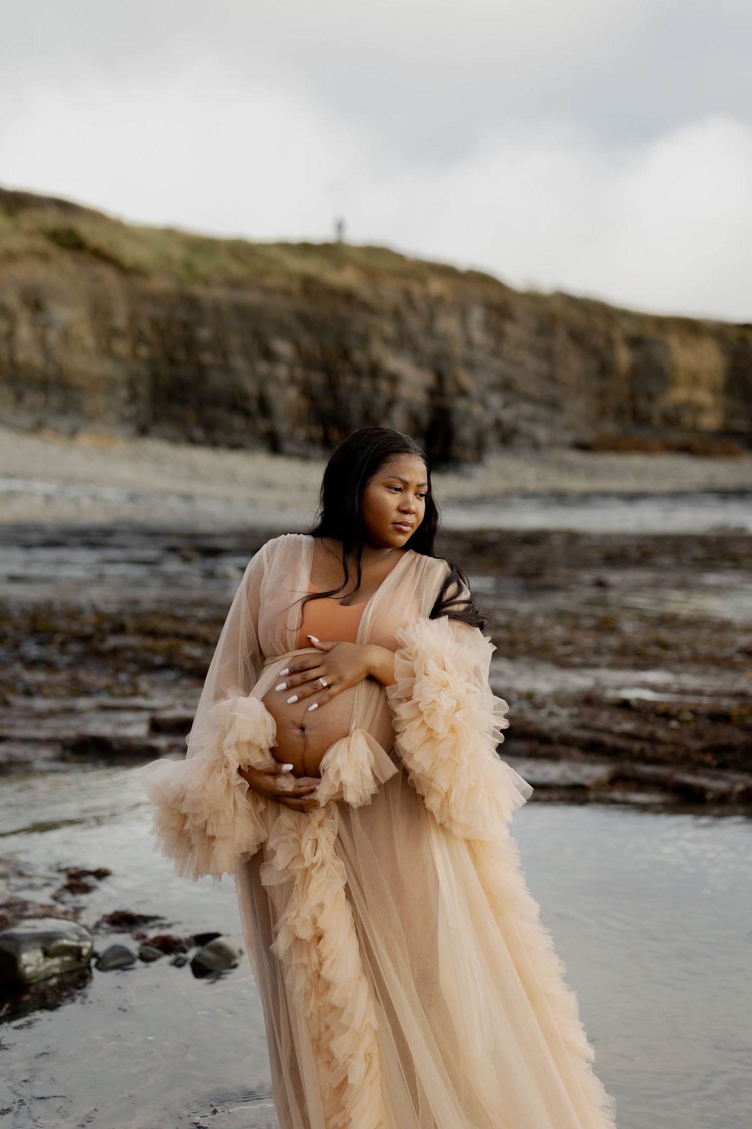 Pregnant woman in flowing beige tulle robe cradling her baby bump on a rocky beach with cliffs in background.