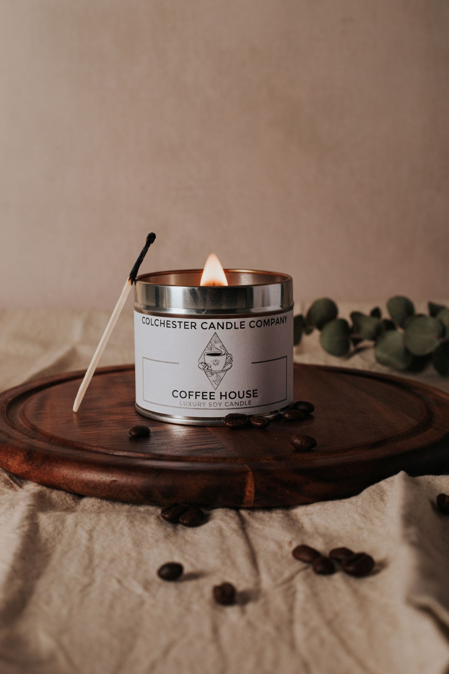 Coffee beans scattered around a white Rochester Candle Company coffee house scented candle on a wooden surface.