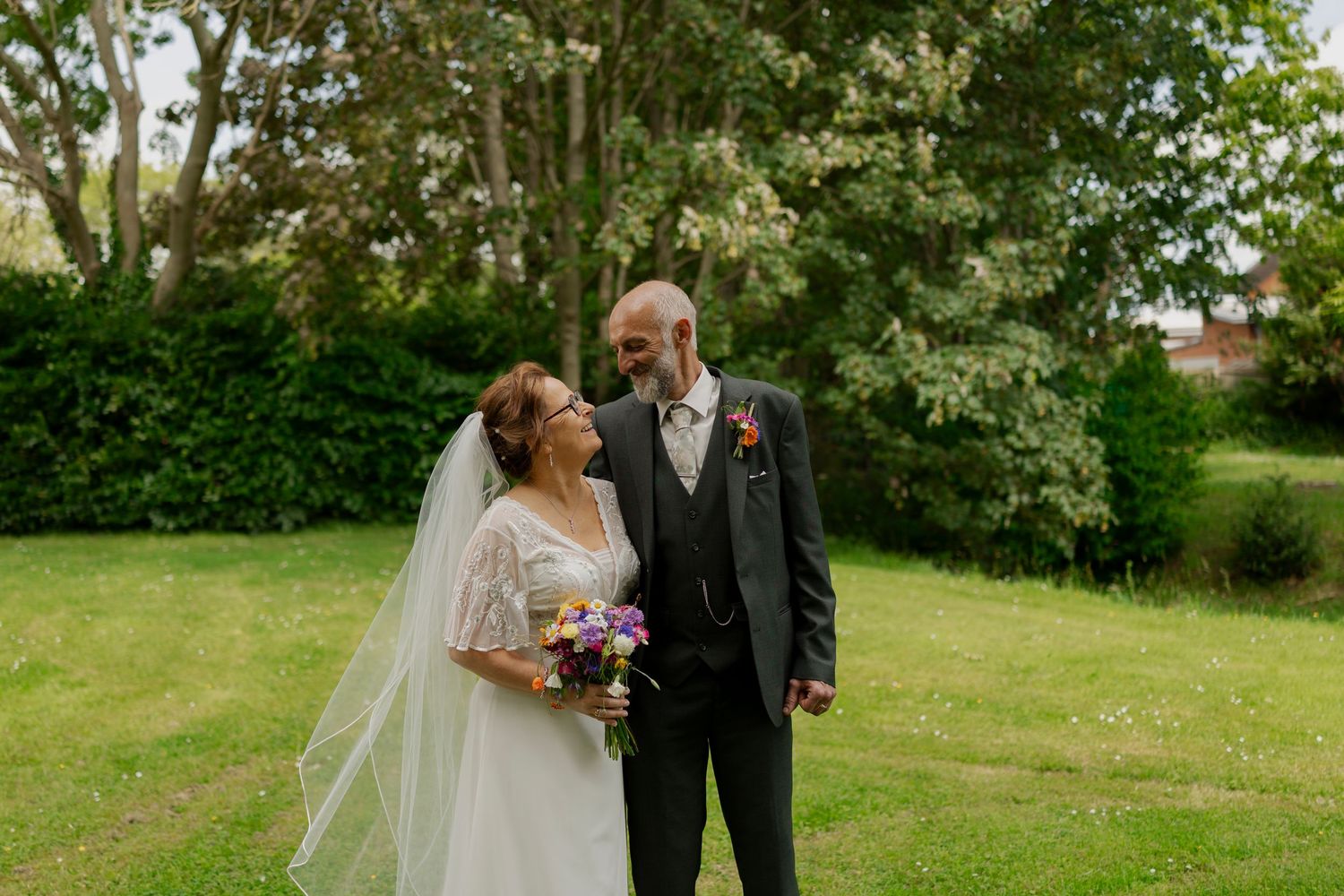 Wedding couple walks hand in hand through a lush green garden on a bright sunny day.