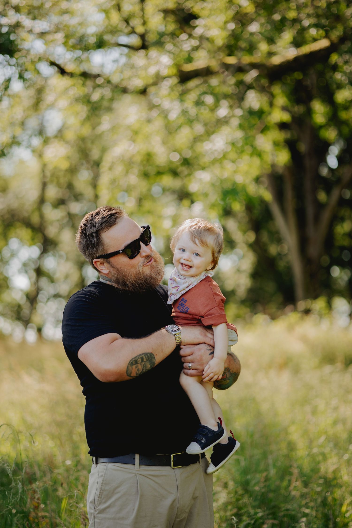 Parent lifting and playing with child while standing in a sunlit field with trees.