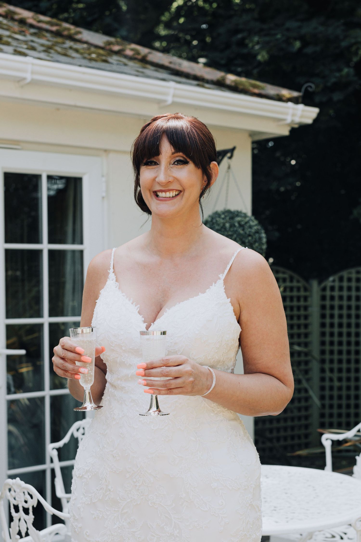 Woman in white lace dress holds champagne glasses at outdoor garden party.