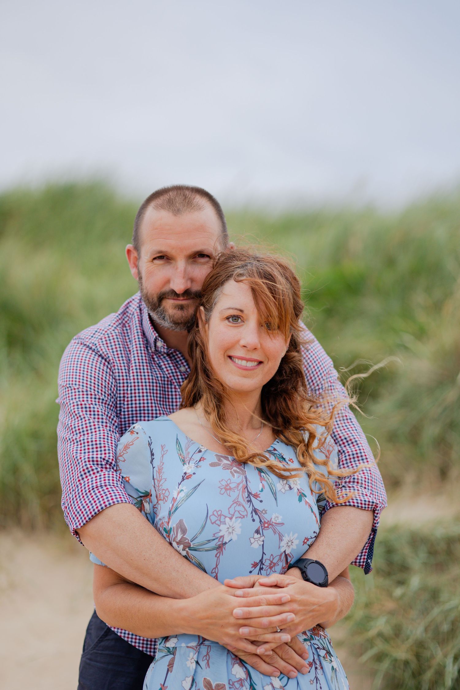 A couple poses for a portrait on a coastal path with beach grass in the background.