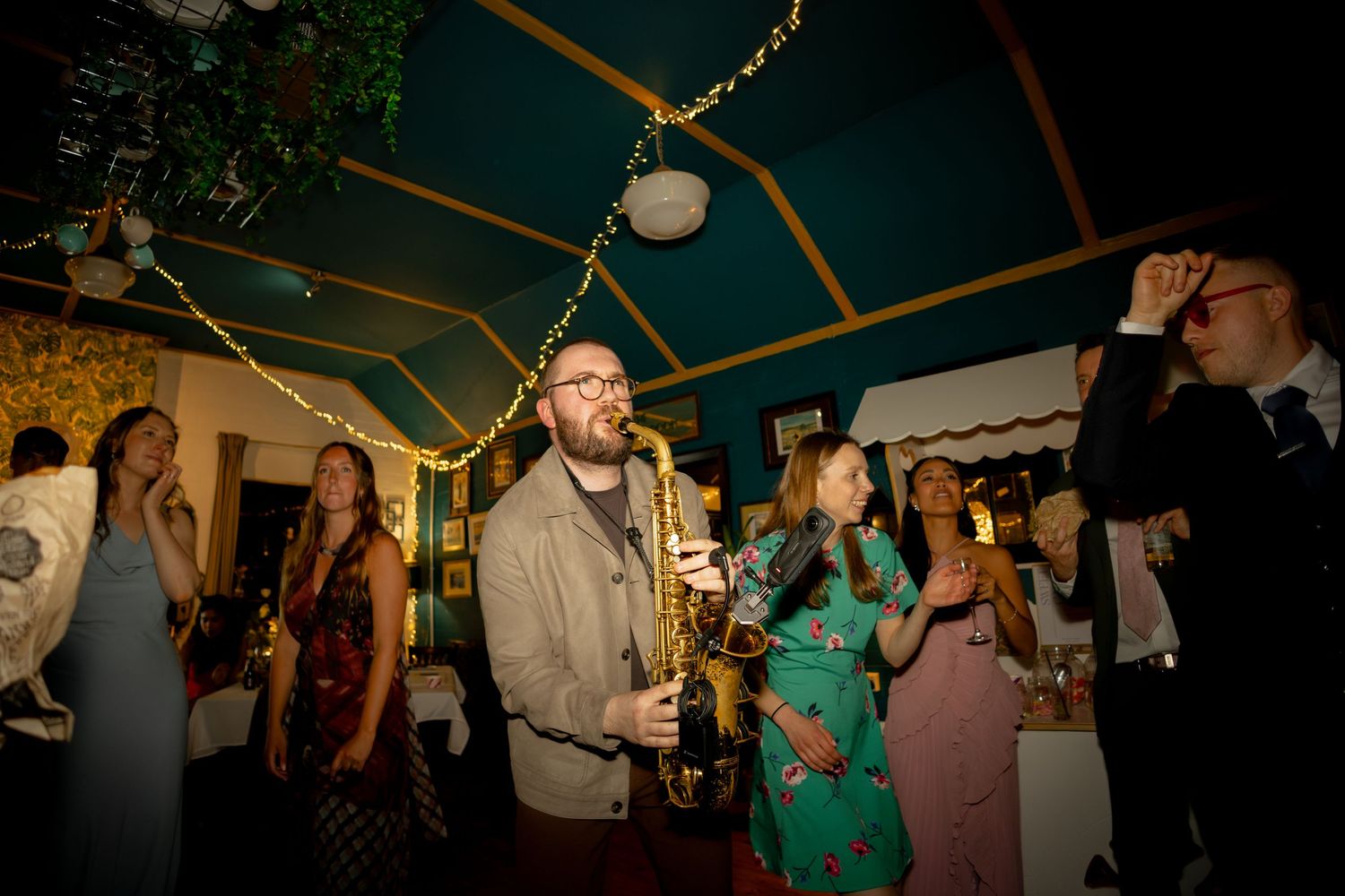Guests socializing at an indoor party under a teal tent with string lights, holding drinks in a warmly lit venue.