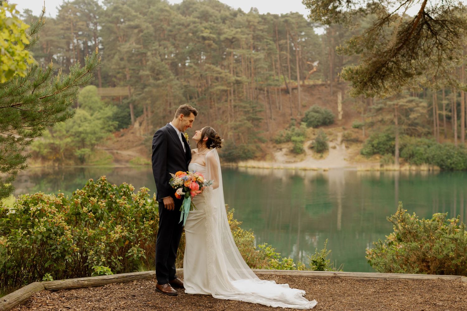 Bride and groom share romantic moment by serene lake surrounded by pine trees, bride holding colorful bouquet in outdoor wedding portrait.