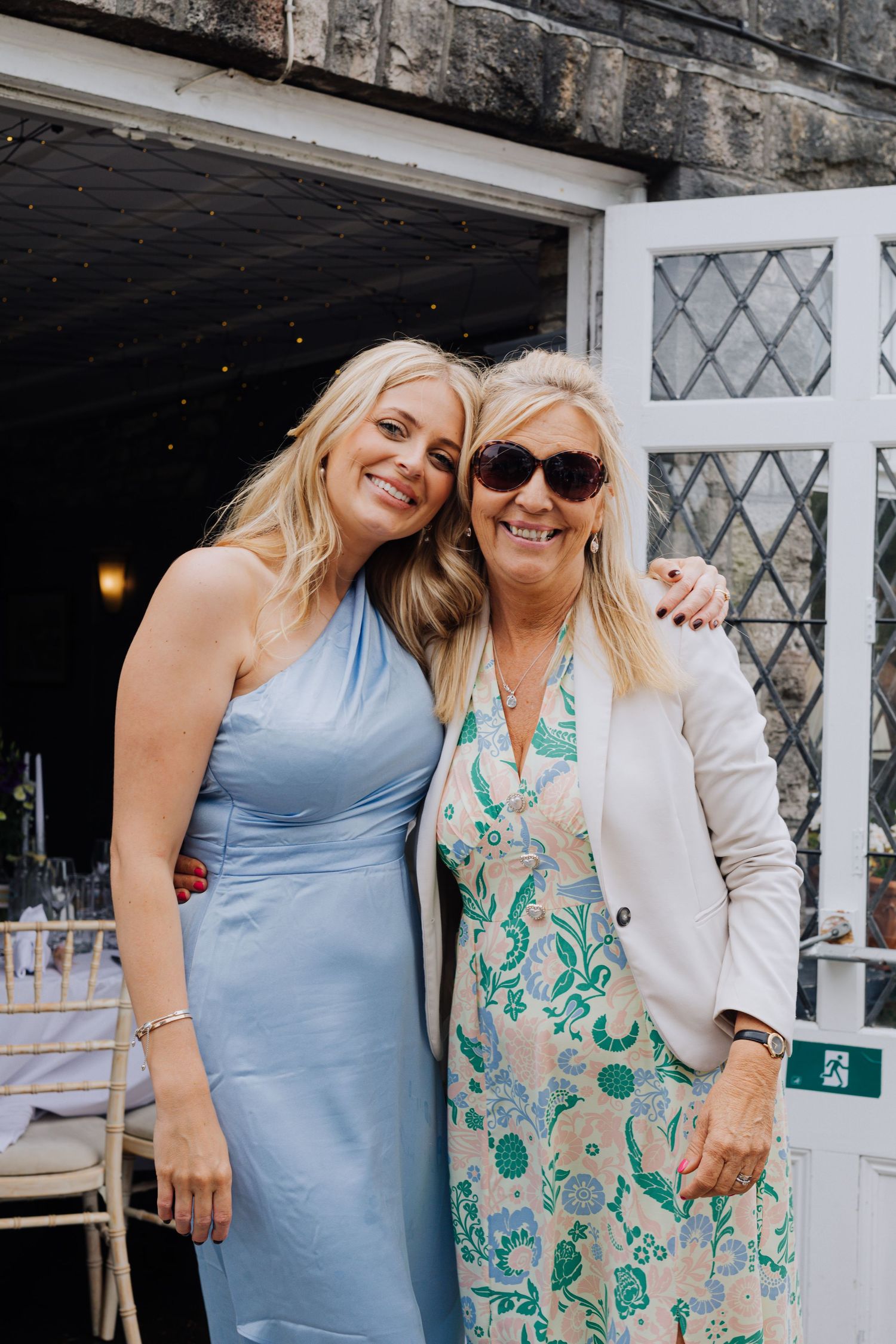 Wedding guests in blue dress and floral print dress pose together at outdoor venue.