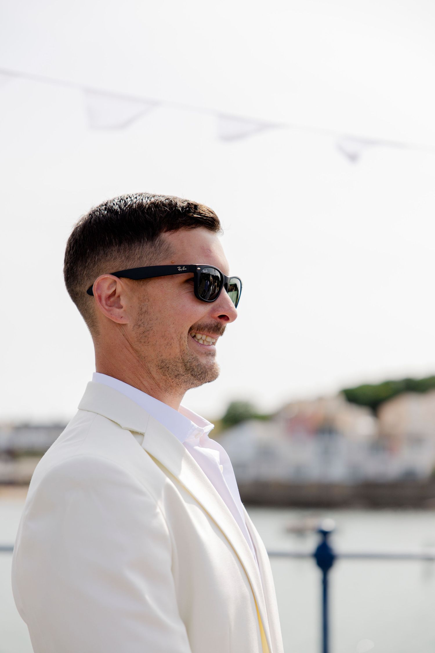 Groom in white suit and sunglasses smiling by waterfront.
