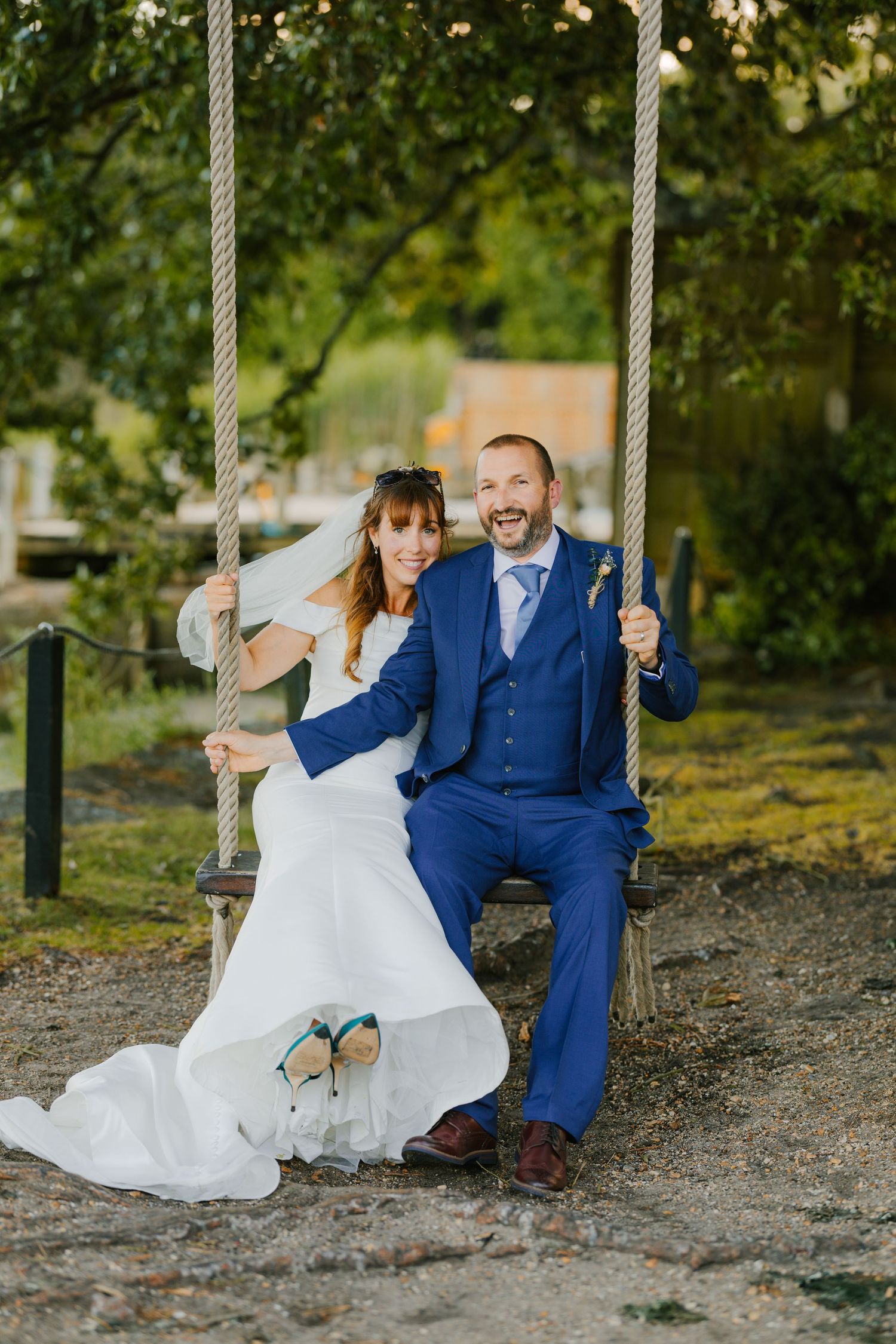 A couple sits together on a swing outdoors, with the bride wearing a white dress and groom in a blue suit.