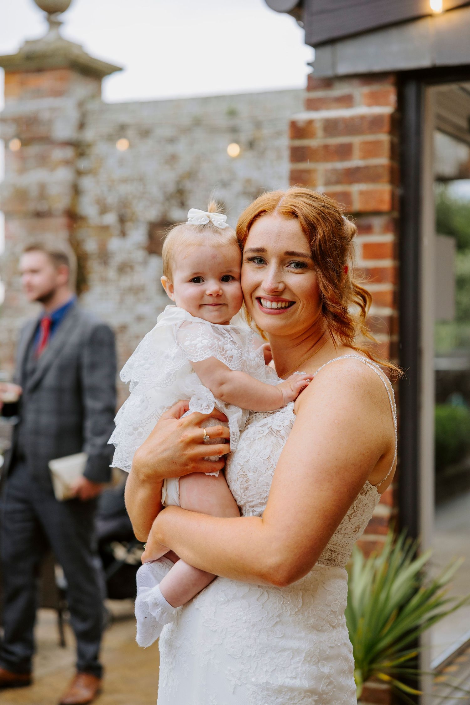 A bride in a white dress holds a small baby wearing a matching white outfit at an outdoor venue.