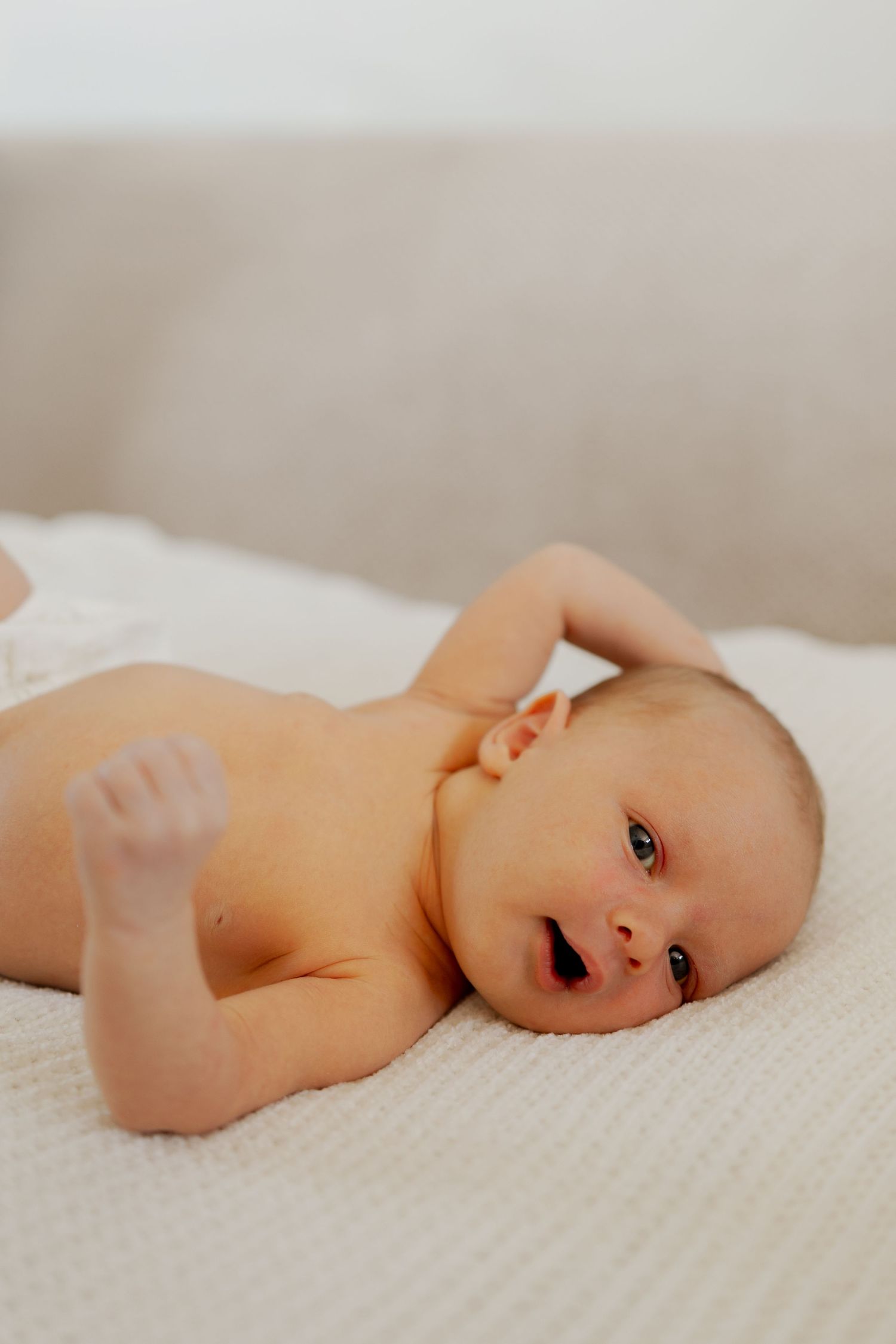 A newborn baby lying on a white textured blanket, with one arm raised, looking alert and calm.