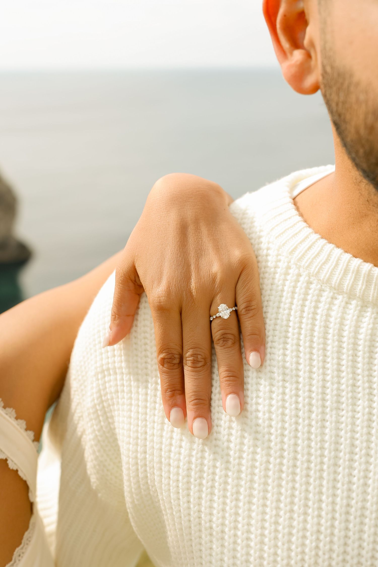 Close-up of a woman's hand displaying a diamond engagement ring resting on a man's white knit sweater shoulder.