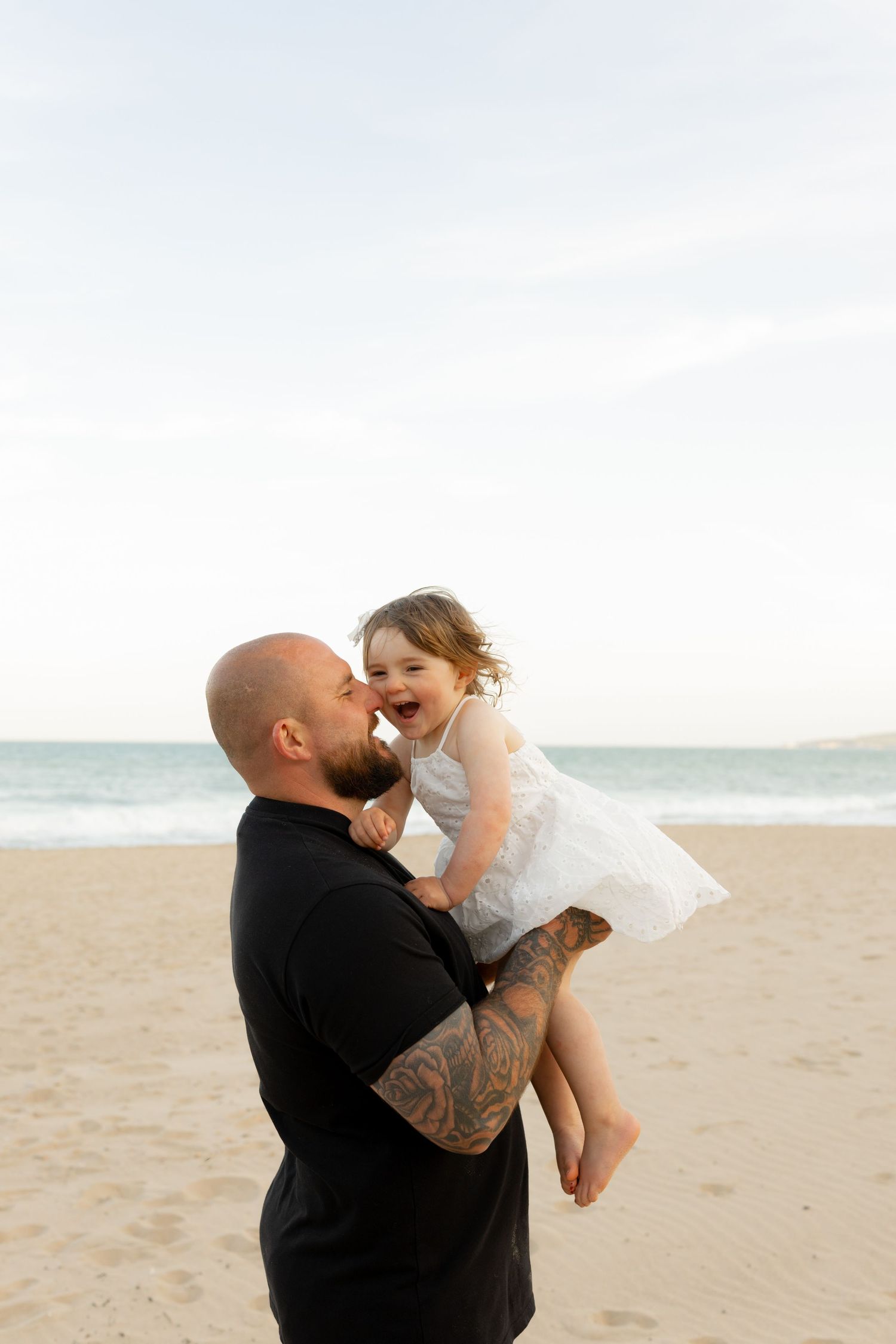 Tattooed father lifting laughing toddler girl in white dress on sandy beach with ocean waves behind them.