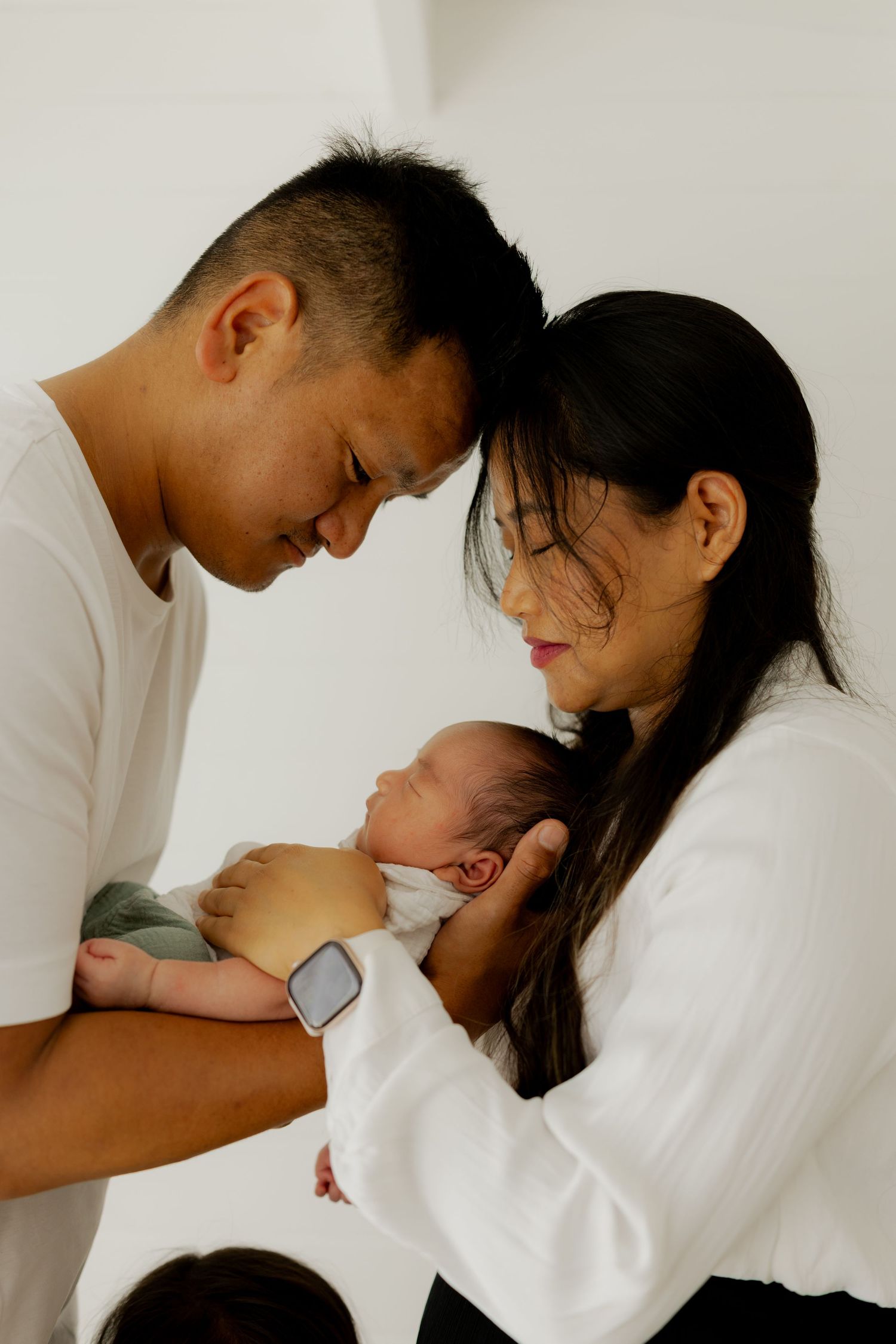 New parents tenderly hold their newborn baby together, foreheads touching in an intimate family portrait.