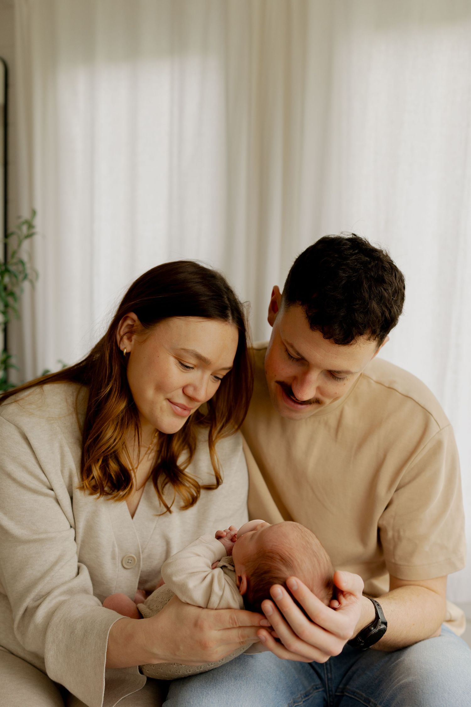 New parents smiling and cradling their sleeping newborn baby together in a bright, cozy room.