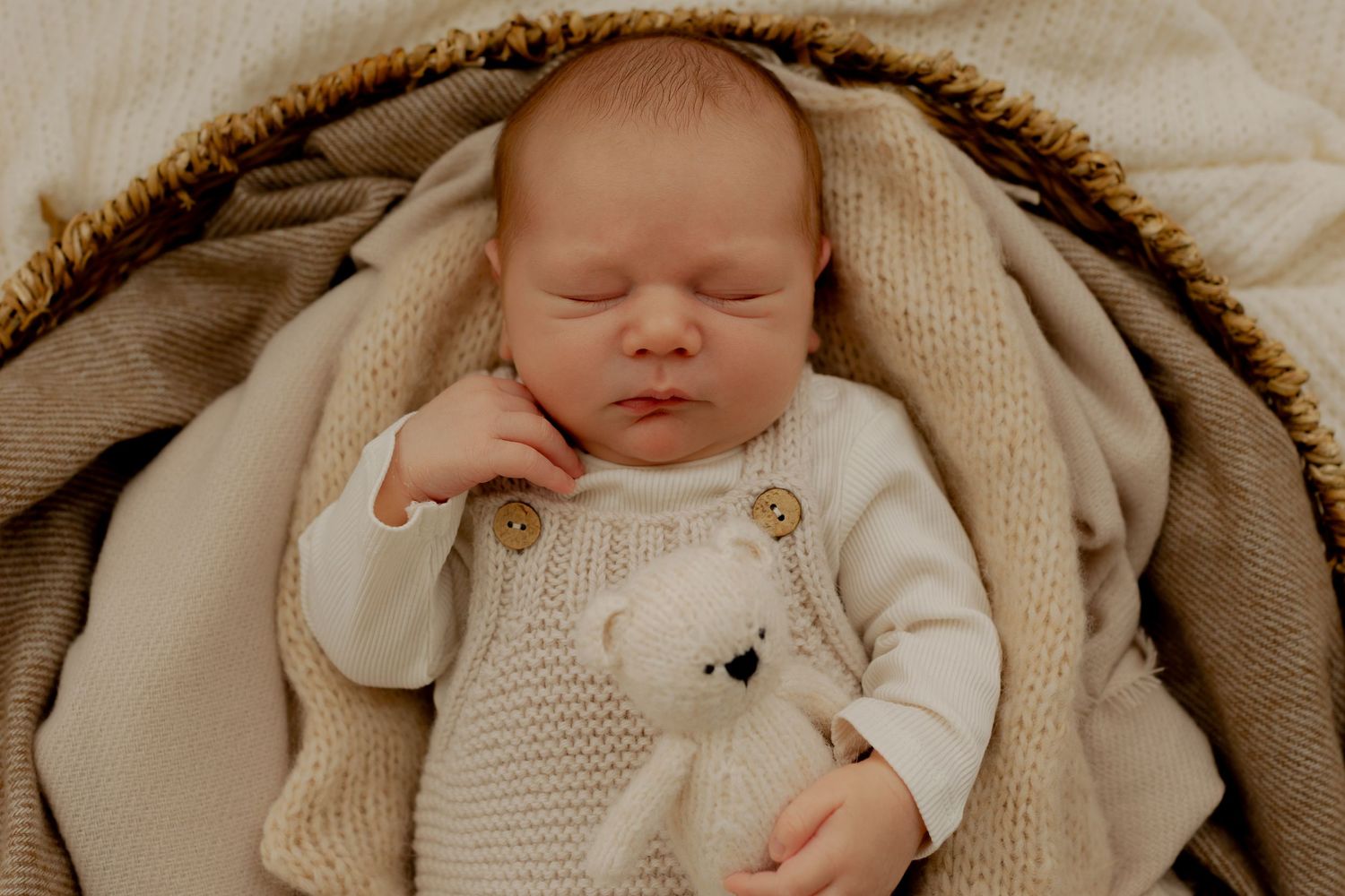 Sleeping newborn baby in knit white bear outfit nestled in a wicker basket with soft beige blankets.