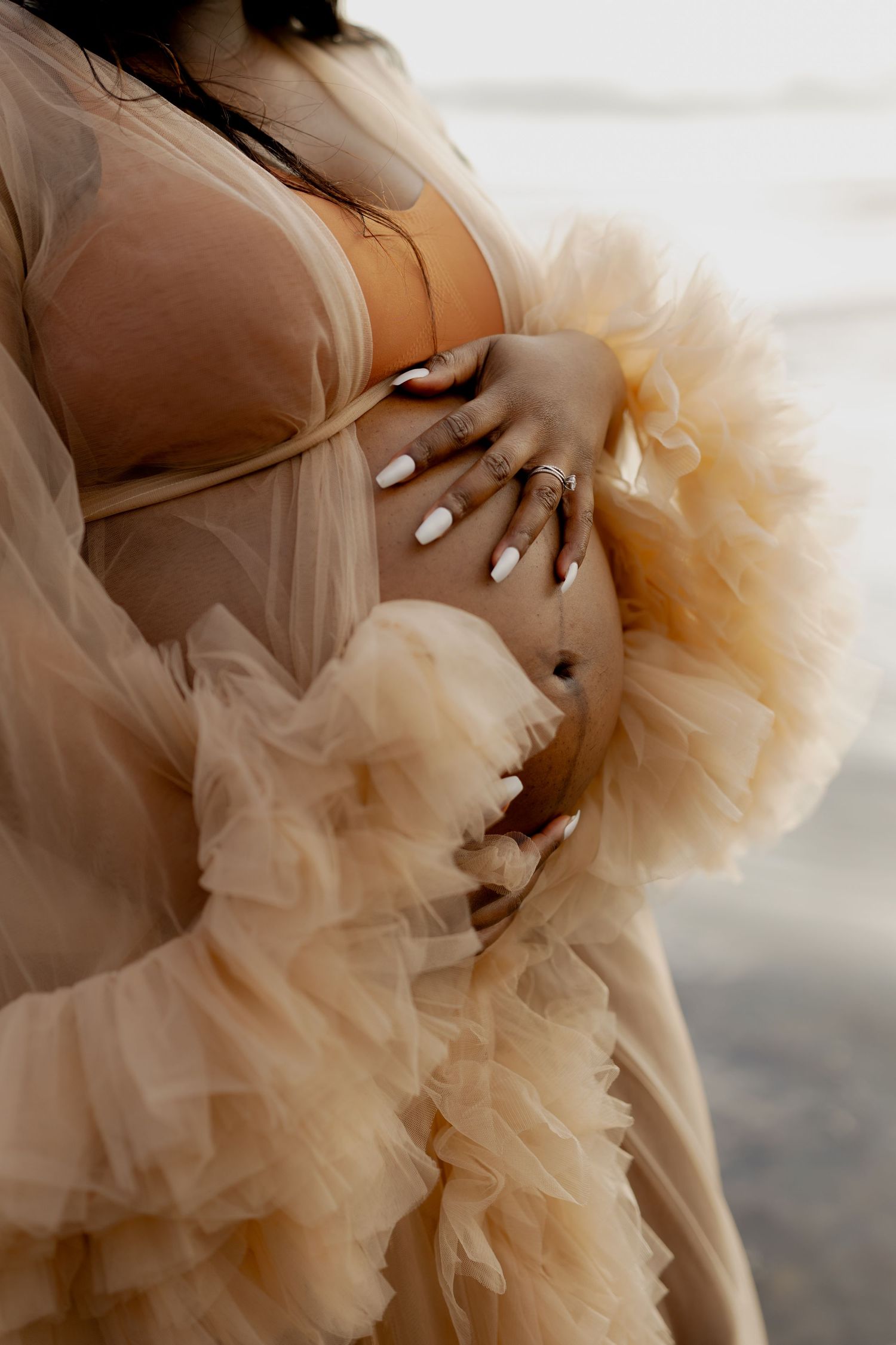 Pregnant woman in a flowing champagne tulle robe cradling her baby bump, wearing white manicured nails and gold jewelry.