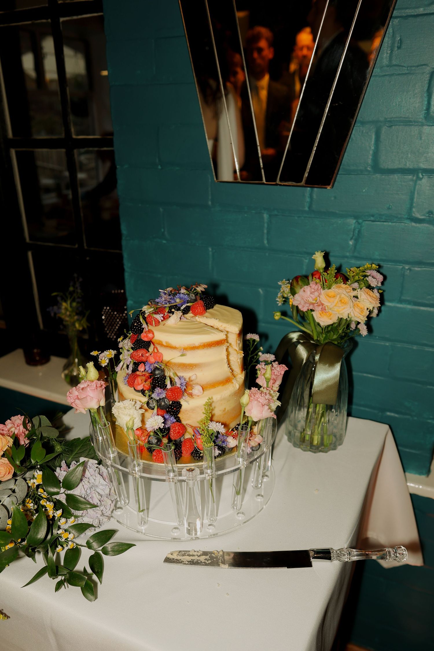 Elegant semi-naked wedding cake adorned with fresh flowers on a crystal stand, set against a teal brick wall backdrop.