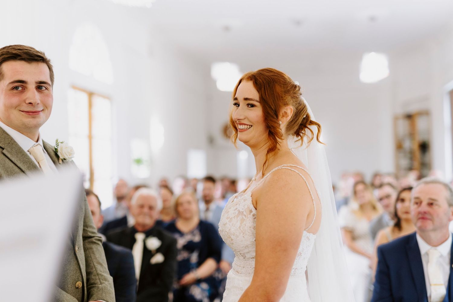 A wedding ceremony in a bright white room with guests watching as the couple exchanges vows at the altar.