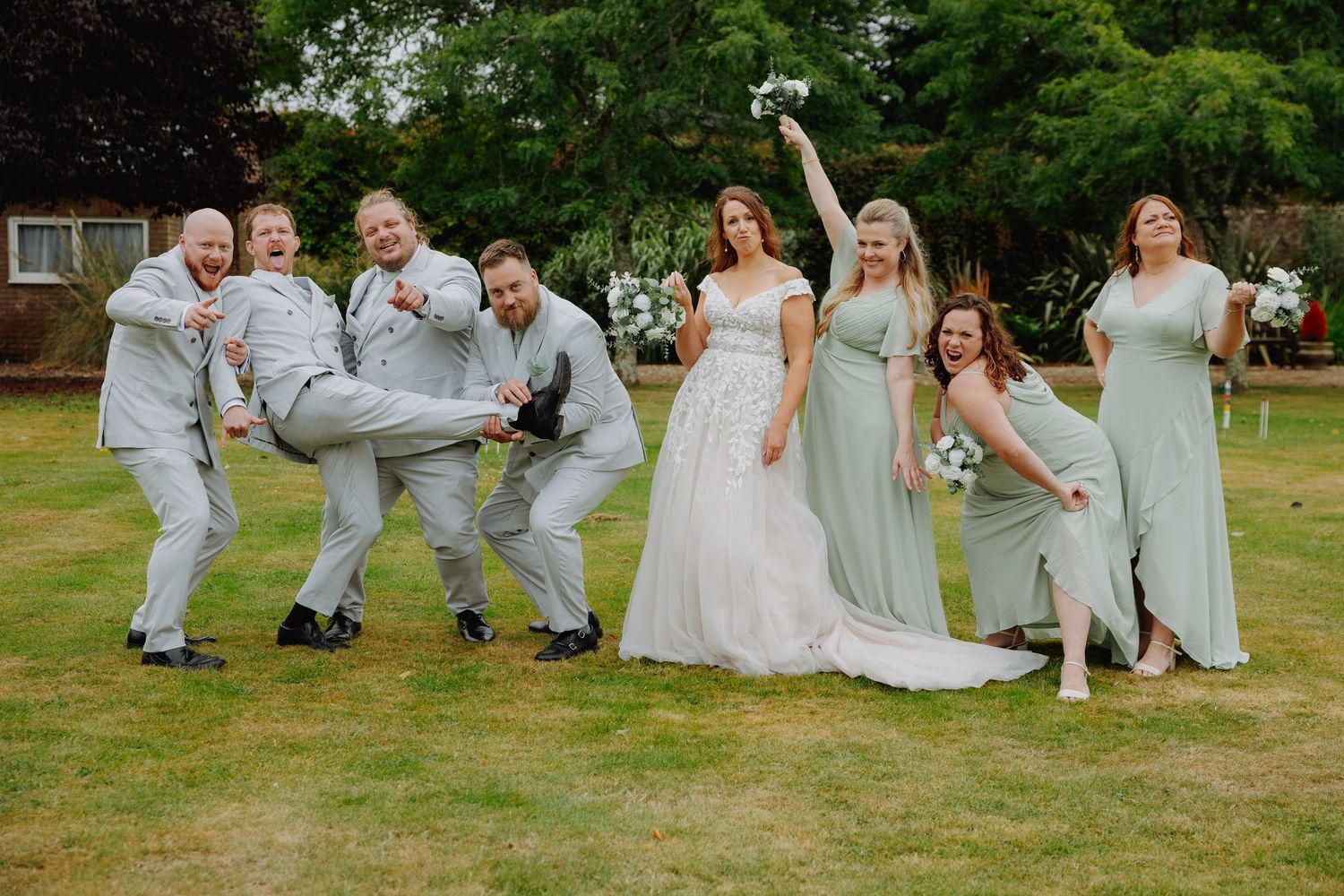 Wedding party in light gray and mint green attire performs fun jumping poses on grassy lawn during outdoor celebration.