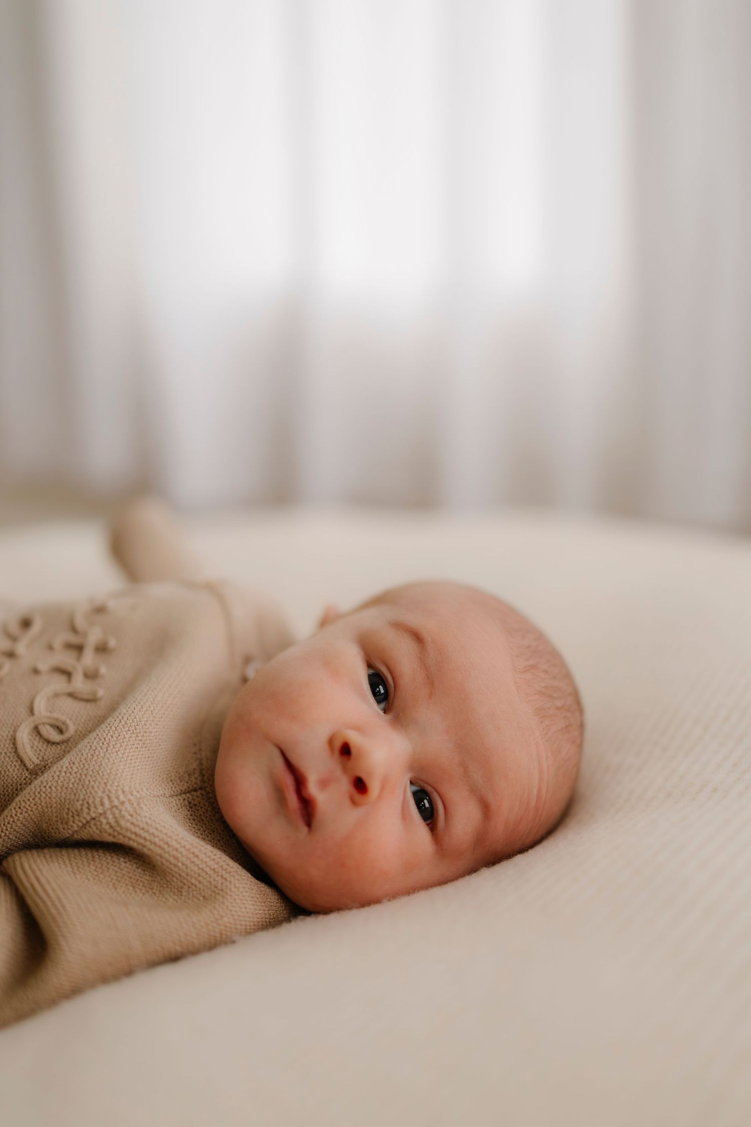 Close-up portrait series of a newborn baby in soft beige sweater lying on white bedding with dreamy lighting.