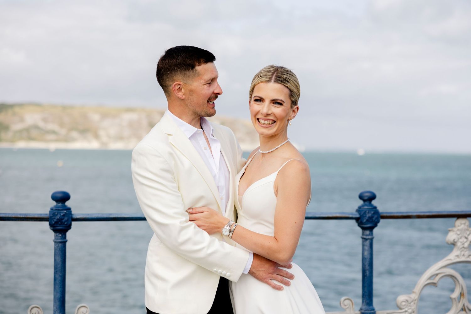 A romantic wedding couple embraces along a seaside railing with ocean views in the background.