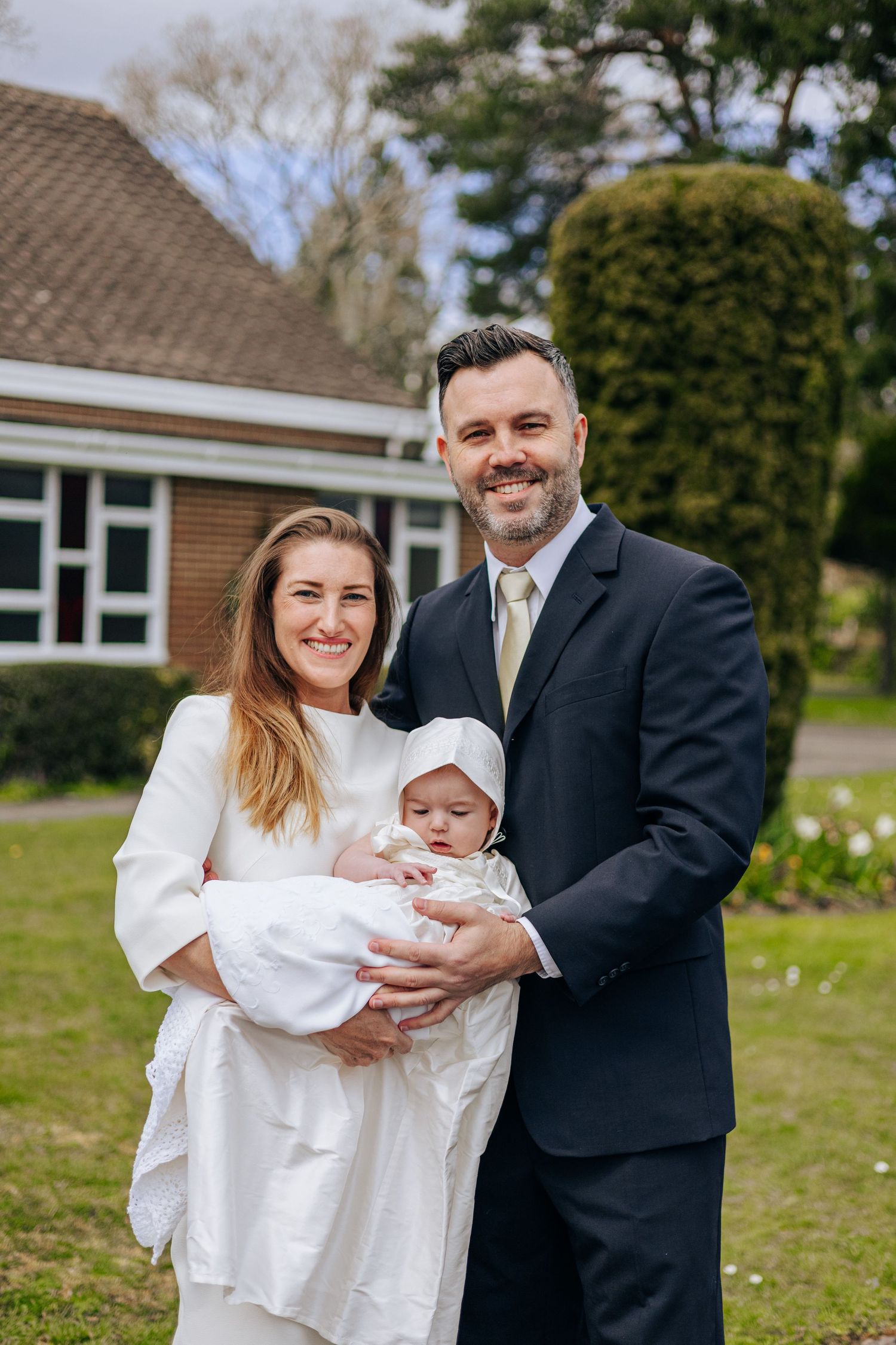 Family dressed in formal attire standing together on grass with baby in white christening gown.