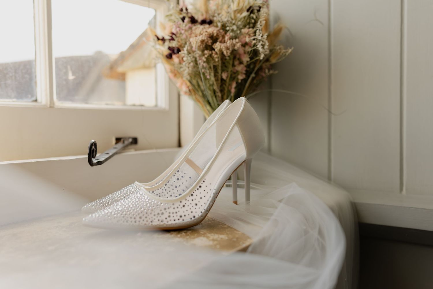 Elegant white crystal-embellished bridal heels resting on a bathtub edge with a dried floral bouquet nearby.