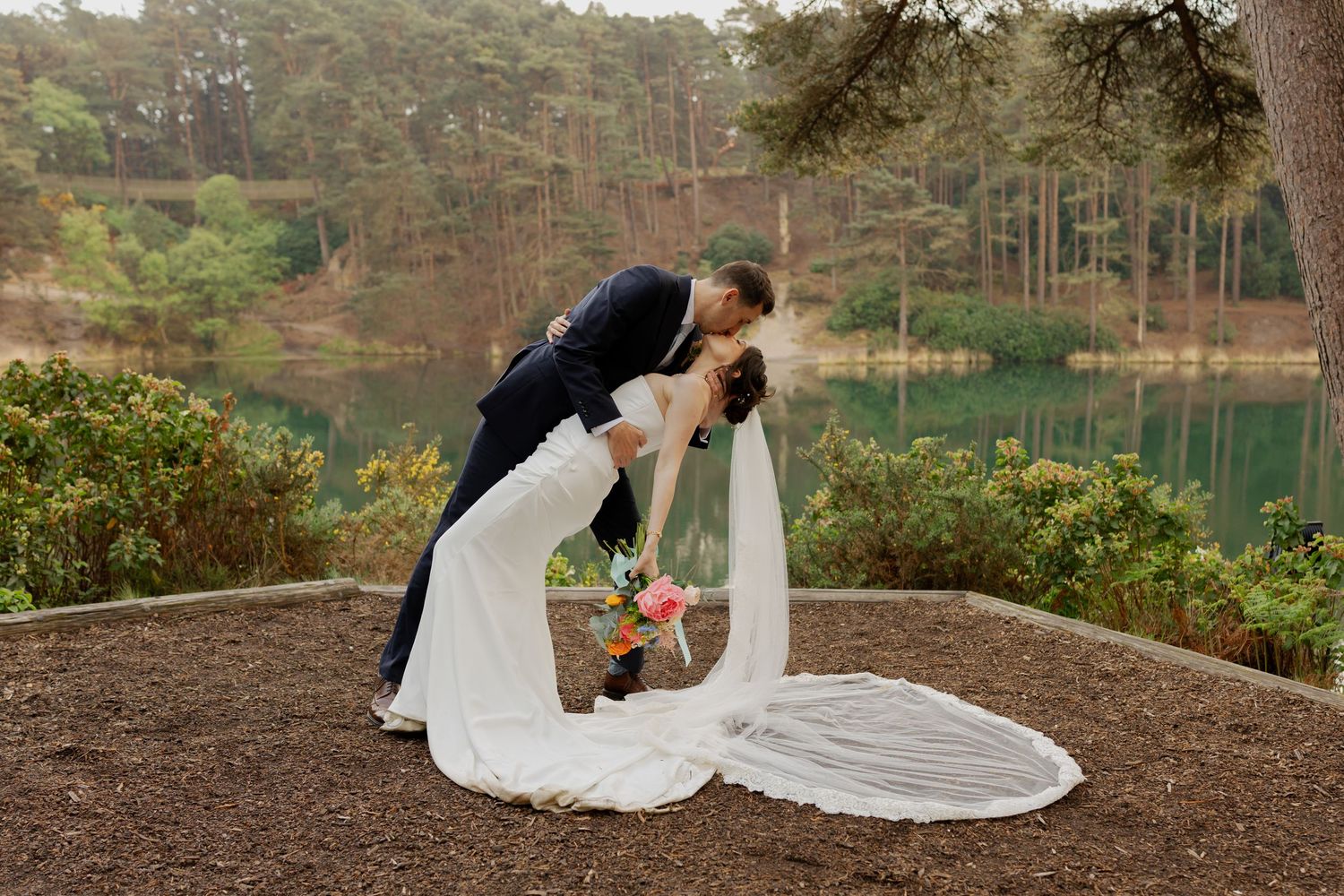 Bride and groom share a romantic dip kiss outdoors near a serene lake, surrounded by lush pine trees.