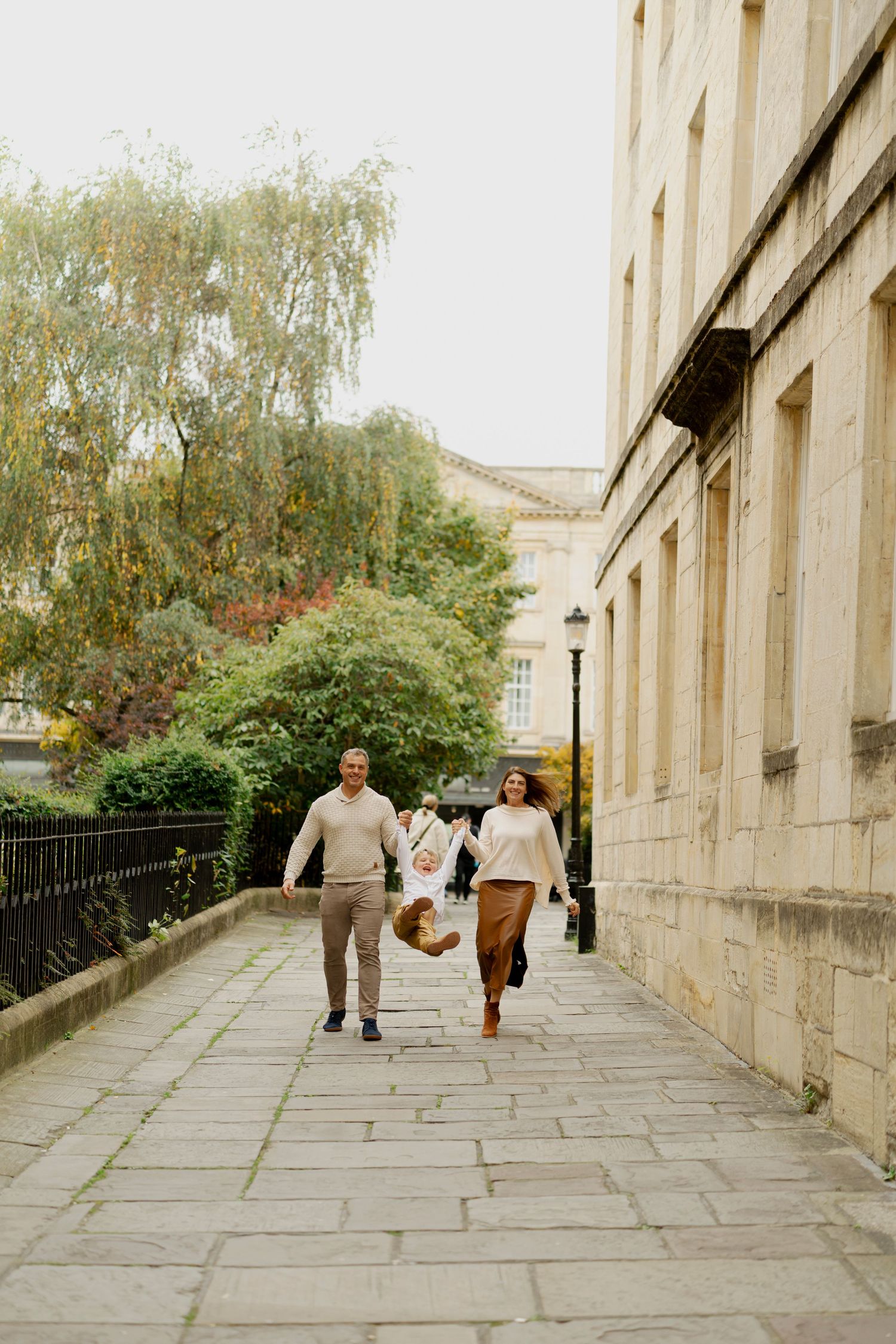 Family of three walking on a stone pathway, parents swinging child between them, beside a classic stone building with lush green trees.