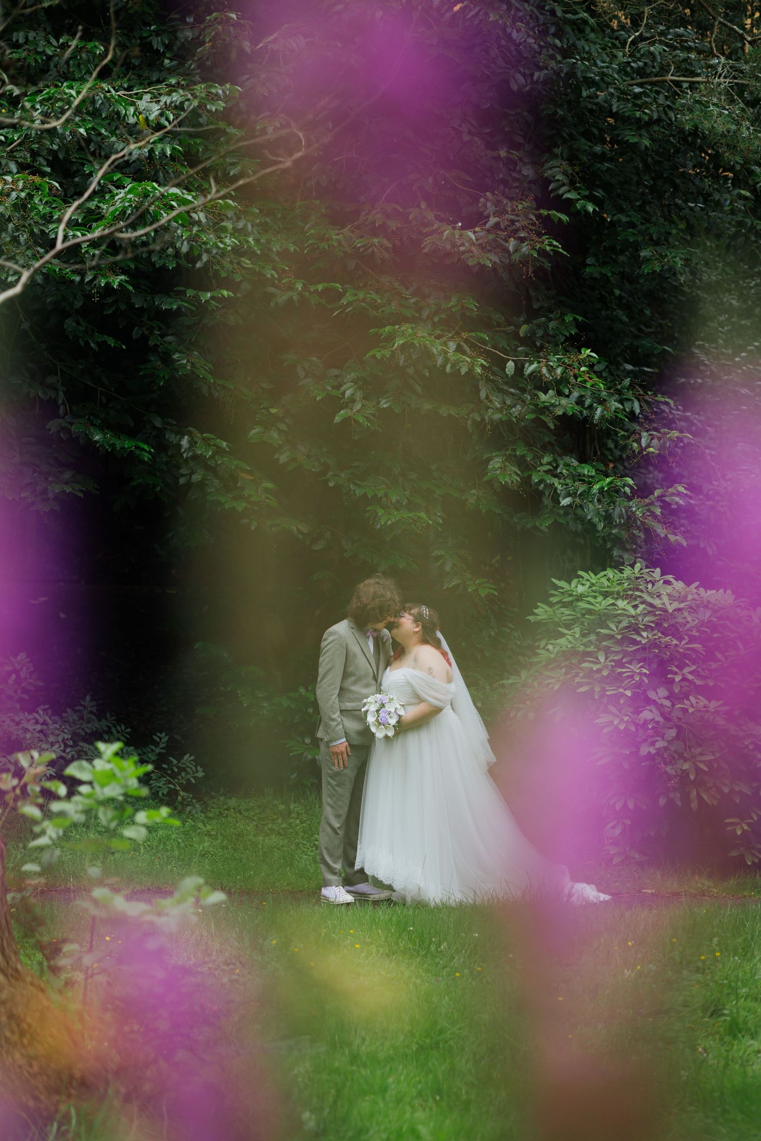 A romantic wedding couple shares an intimate moment in a garden setting framed by purple flowers and greenery.
