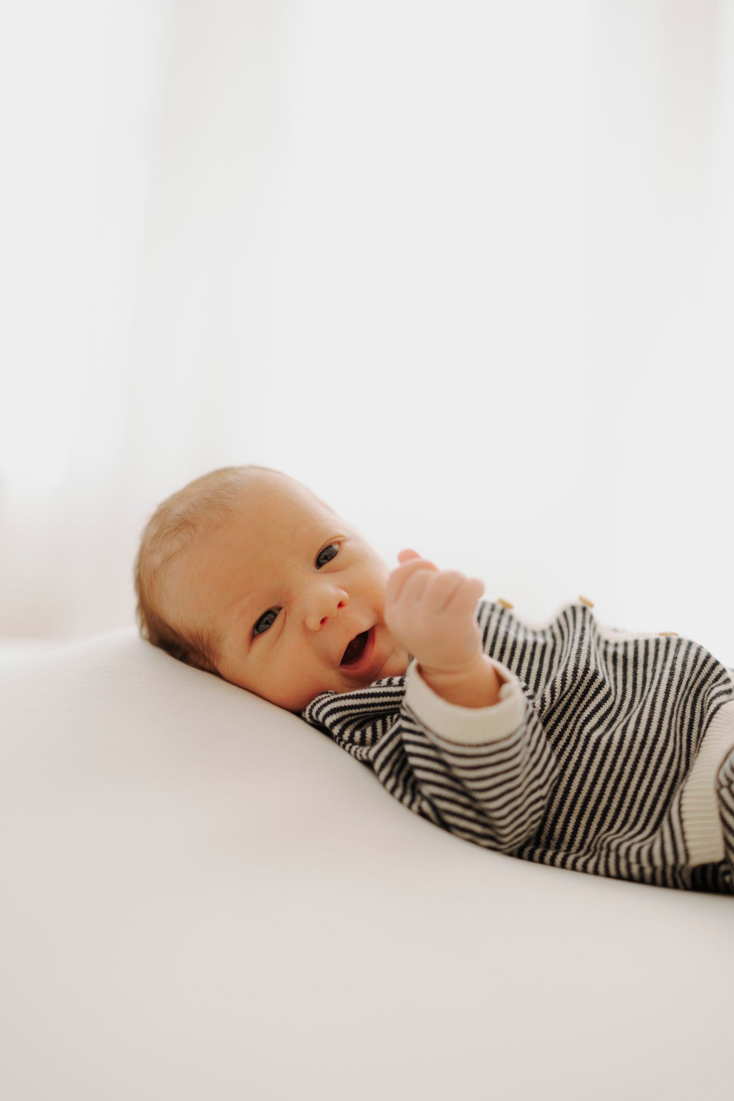 Baby wearing striped onesie lying on white bedding with a sweet expression.