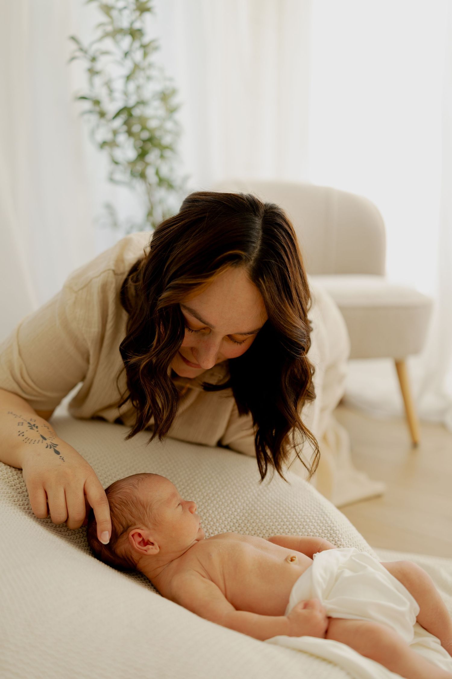 Parent lovingly kisses sleeping baby while resting on textured white bedding in bright minimalist room.
