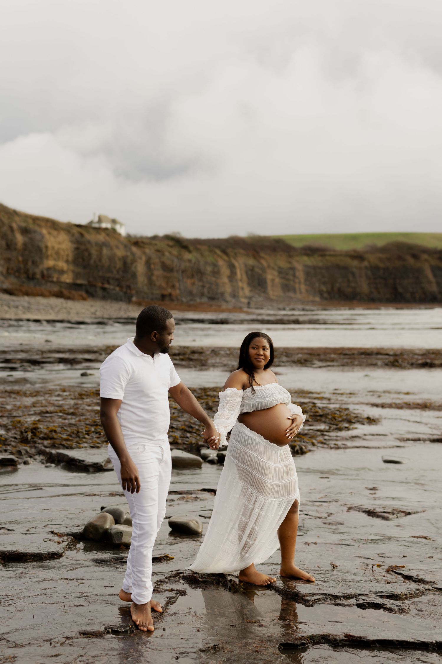 A pregnant couple in white outfits holds hands on a rocky beach with dramatic cliffs in the background.