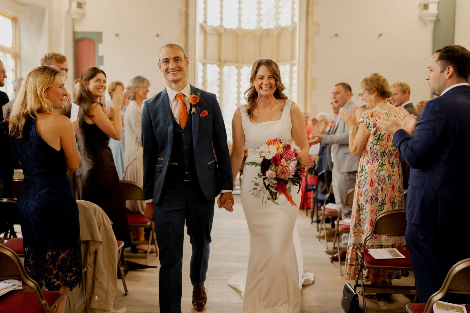 Two people walk down the aisle at a wedding ceremony while guests stand and cheer in a sunlit indoor venue.