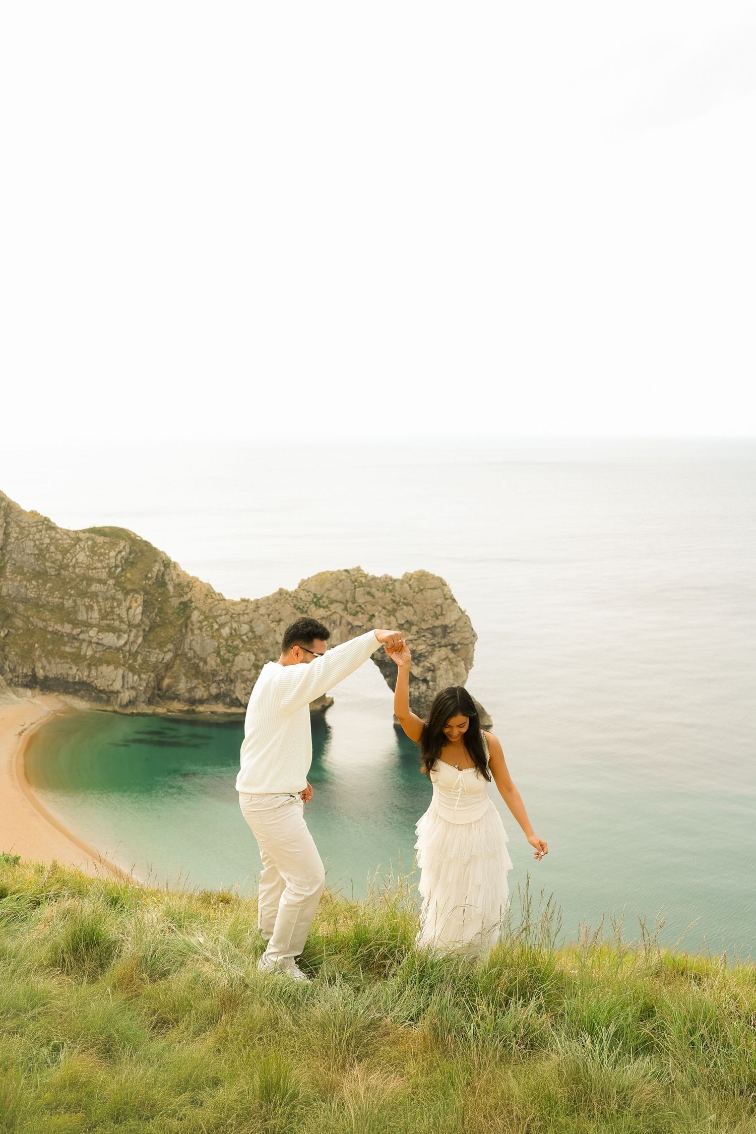 Couple in white attire dancing on a cliff overlooking Durdle Door arch and turquoise sea in Dorset.