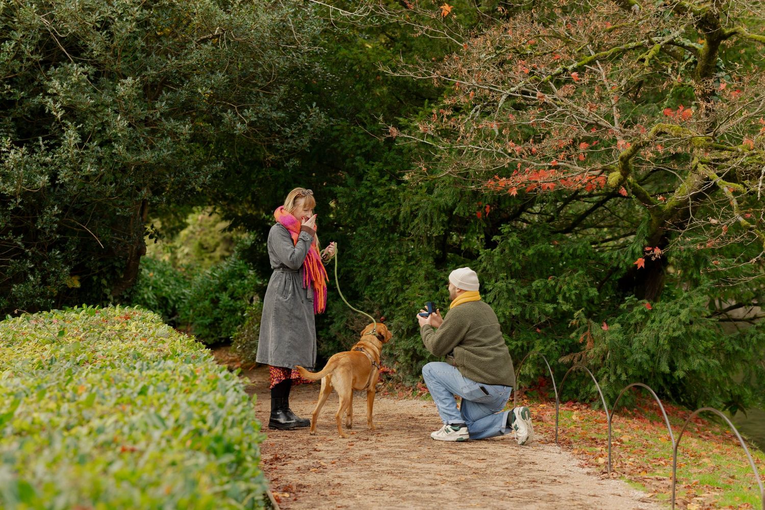 Two people with a brown dog on a garden path surrounded by lush greenery and autumn foliage.