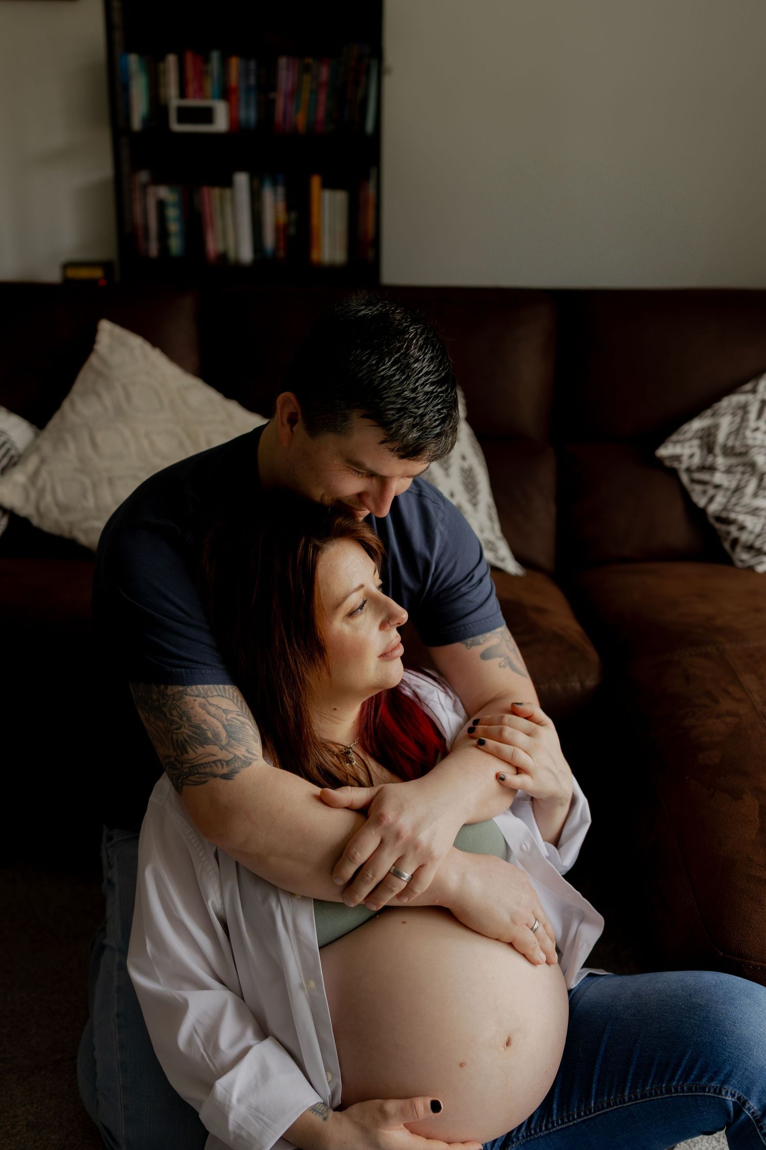 A couple shares an intimate moment during a maternity photoshoot in a dimly lit room with bookshelves in the background.