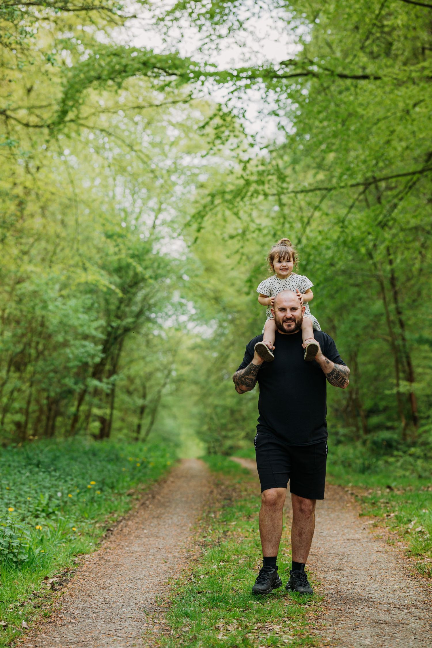 Adult carrying child on shoulders while walking down tree-lined forest path.