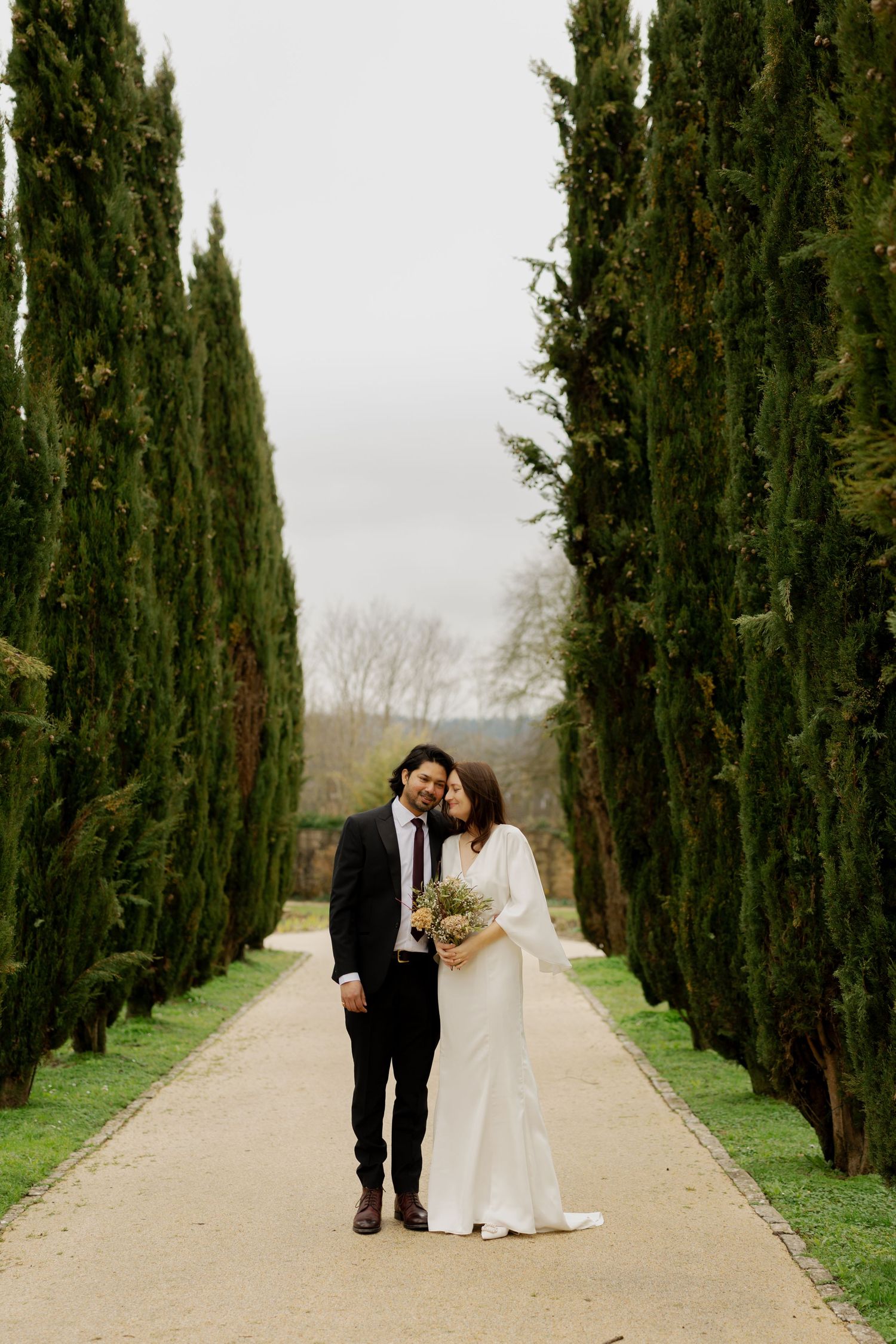 A wedding couple walks down a scenic cypress tree-lined path in the countryside.