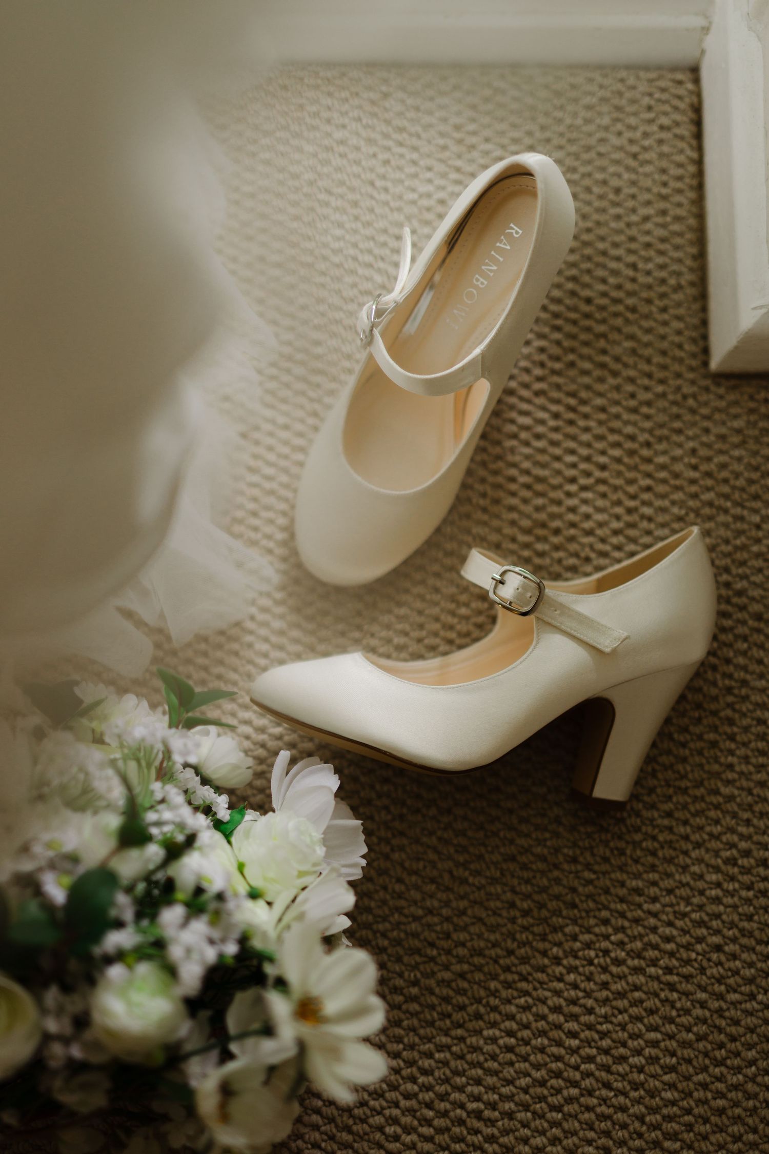 Elegant white bridal heels with ankle straps next to a floral bouquet on beige carpet.
