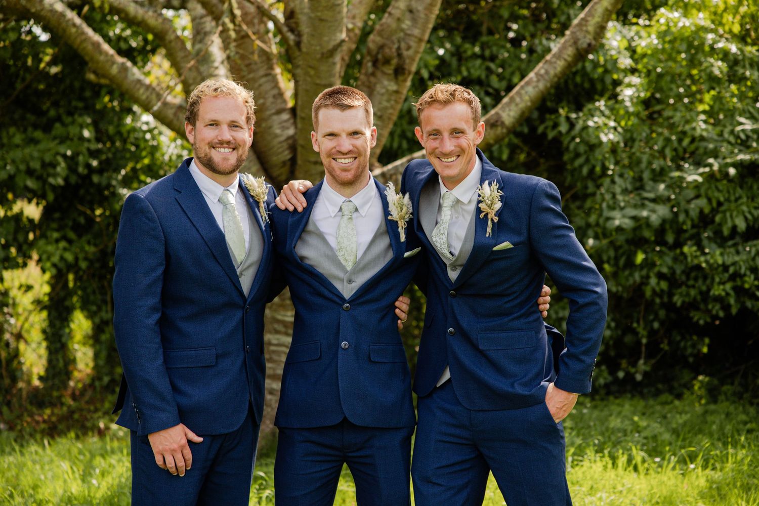 Three groomsmen in matching navy suits pose together on a sunny day in a garden setting.