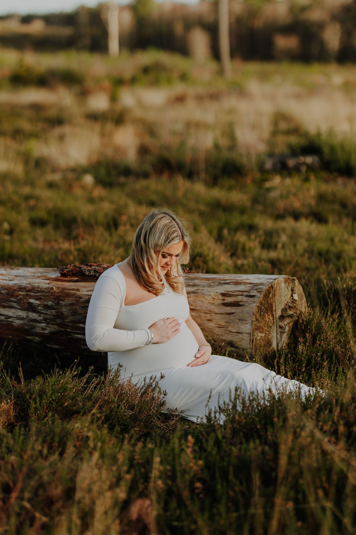 Expectant mother in white dress sitting peacefully on log in golden sunset meadow.