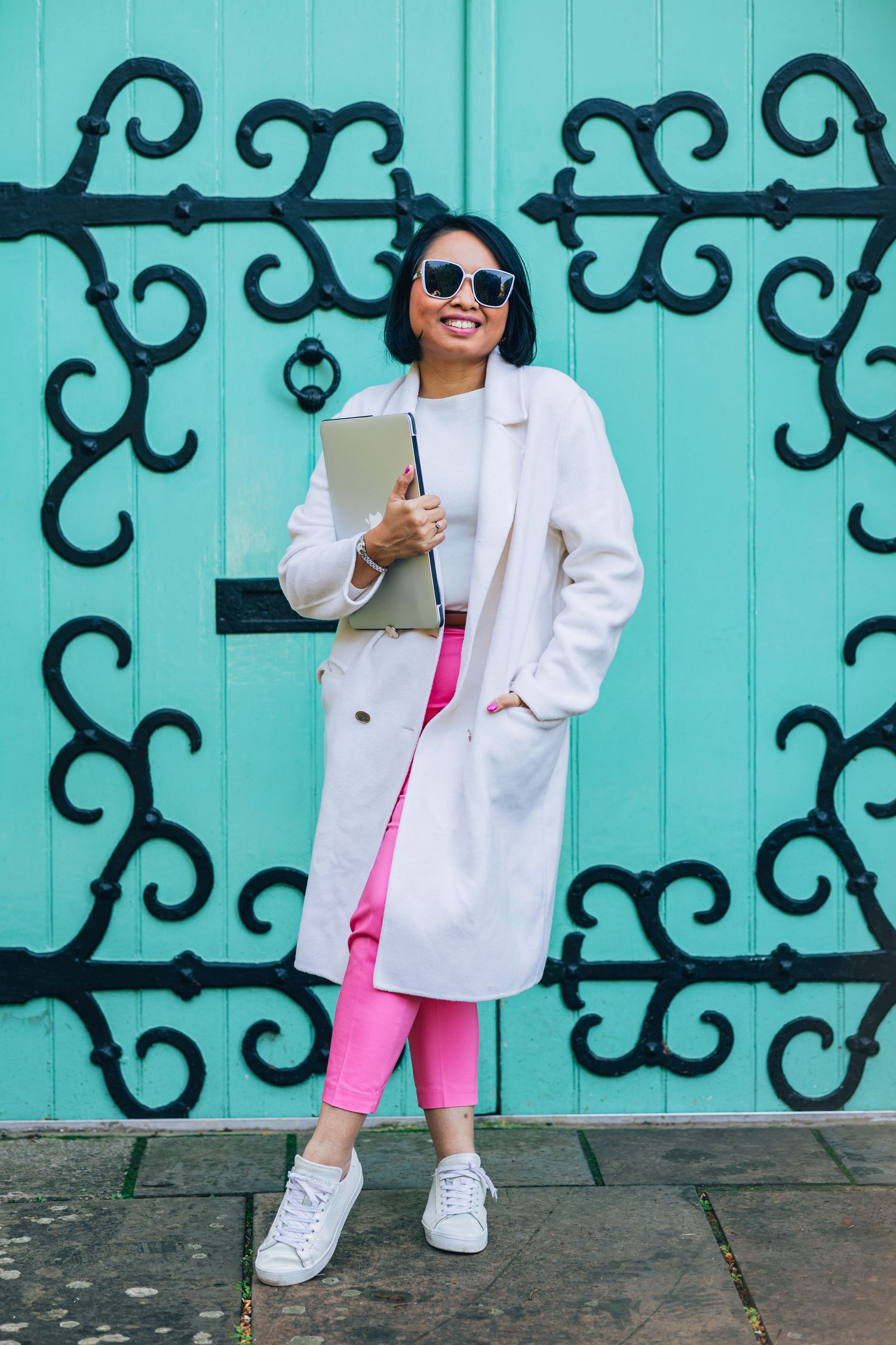 Stylish outfit with white coat and bright pink pants against ornate turquoise door with decorative black ironwork.