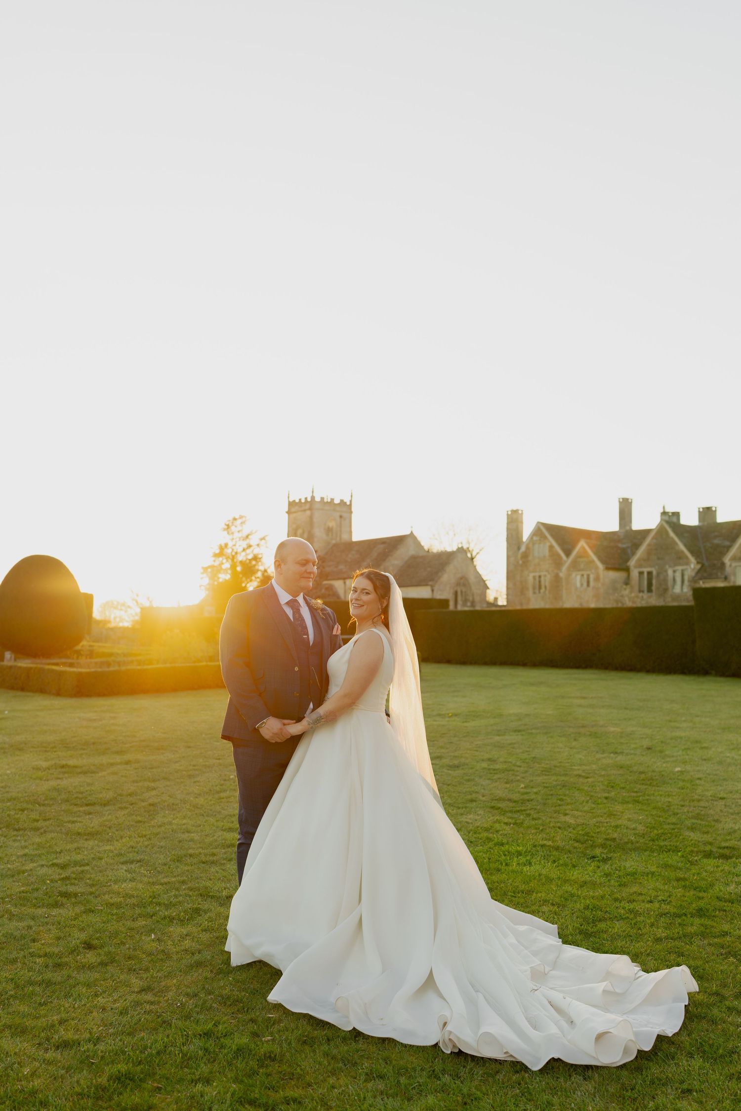 Bride and groom embrace at golden hour sunset with historic English manor and church in background.